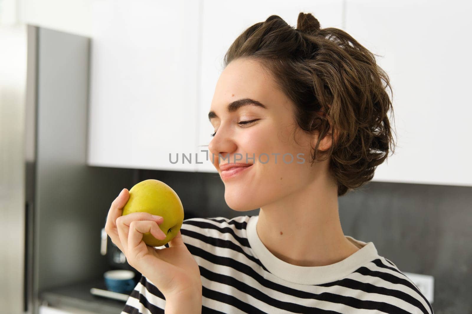 Close up portrait of beautiful, smiling brunette woman, holding an apple with pleased face and closed eyes, standing in the kitchen.