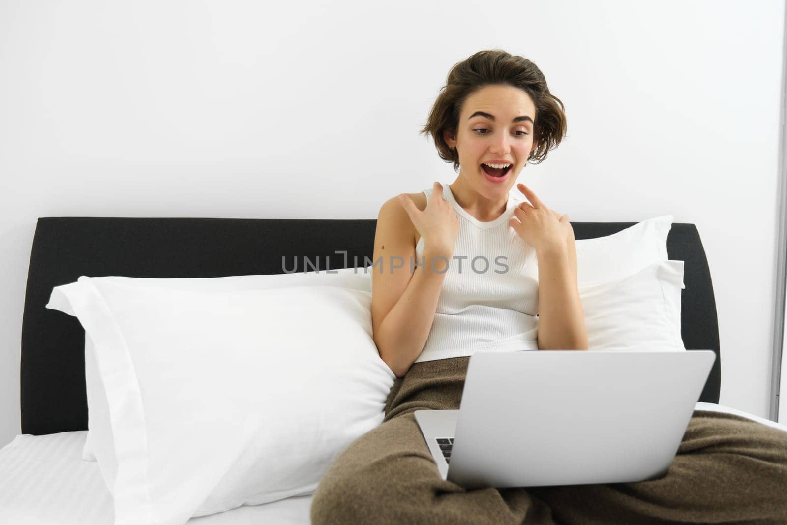 Portrait of woman sitting on bed with laptop, gasping and looking surprised at computer screen. Technology and lifestyle concept