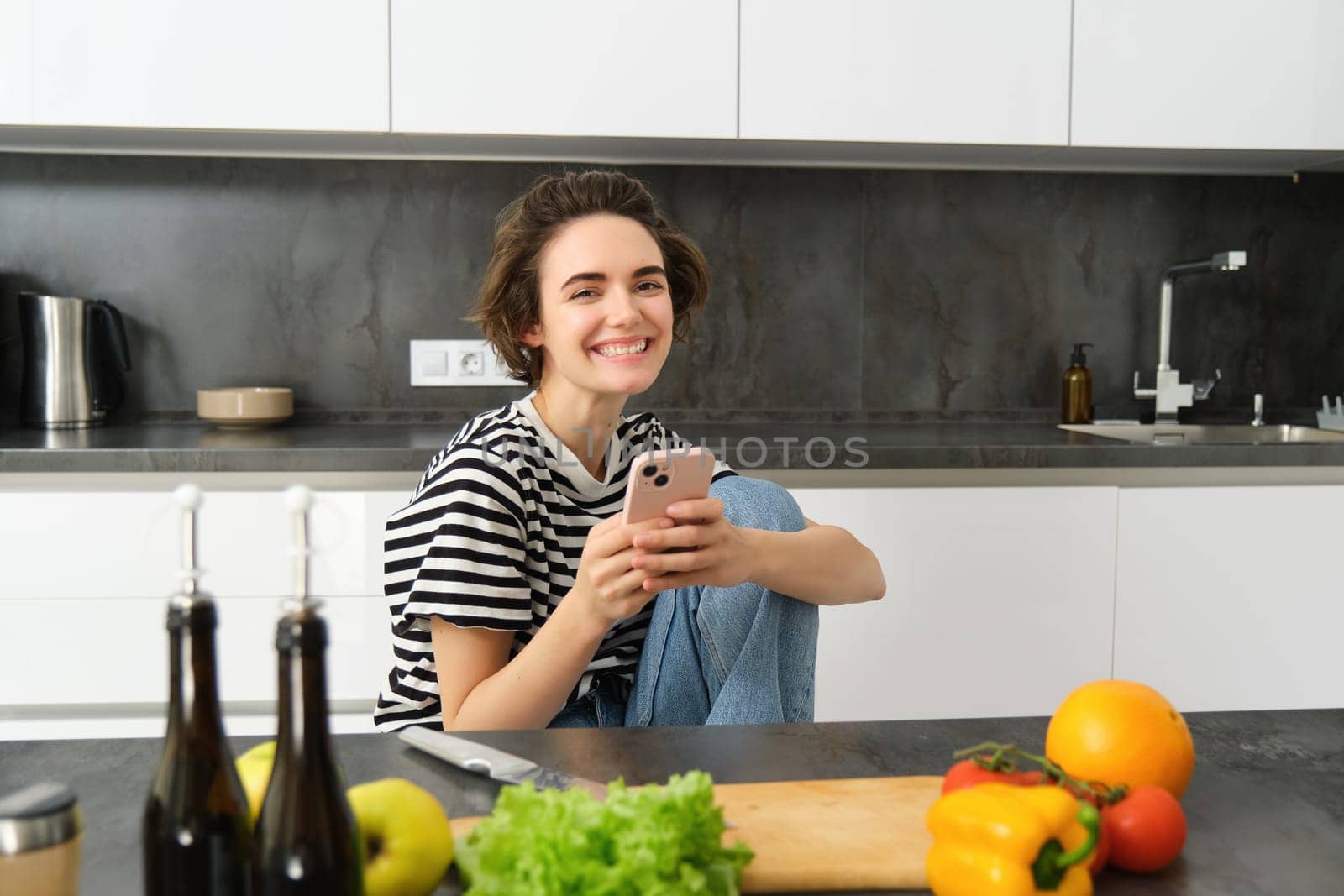 Portrait of young woman searching for cooking recipes online using smartphone, sitting near vegetables, salad ingredients and chopping board, smiling at camera by Benzoix