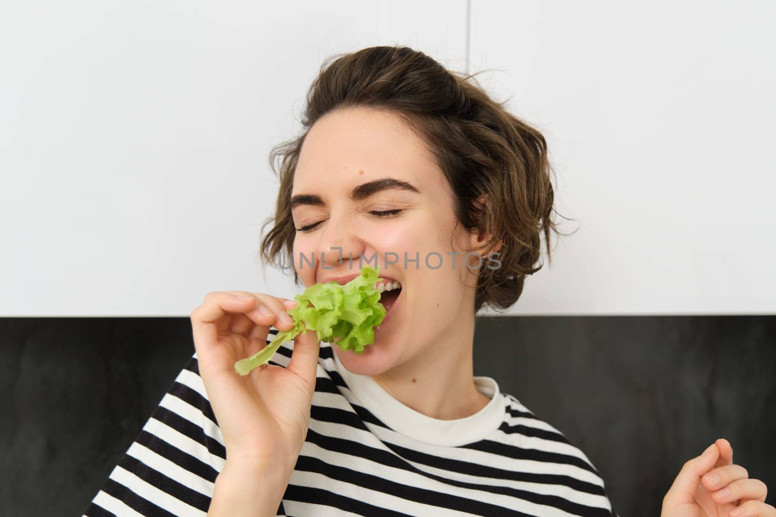 Close up of funny cute woman, vegetarian eating lettuce leaf and smiling, concept of healthy diet, girl likes vegetables, standing in the kitchen.