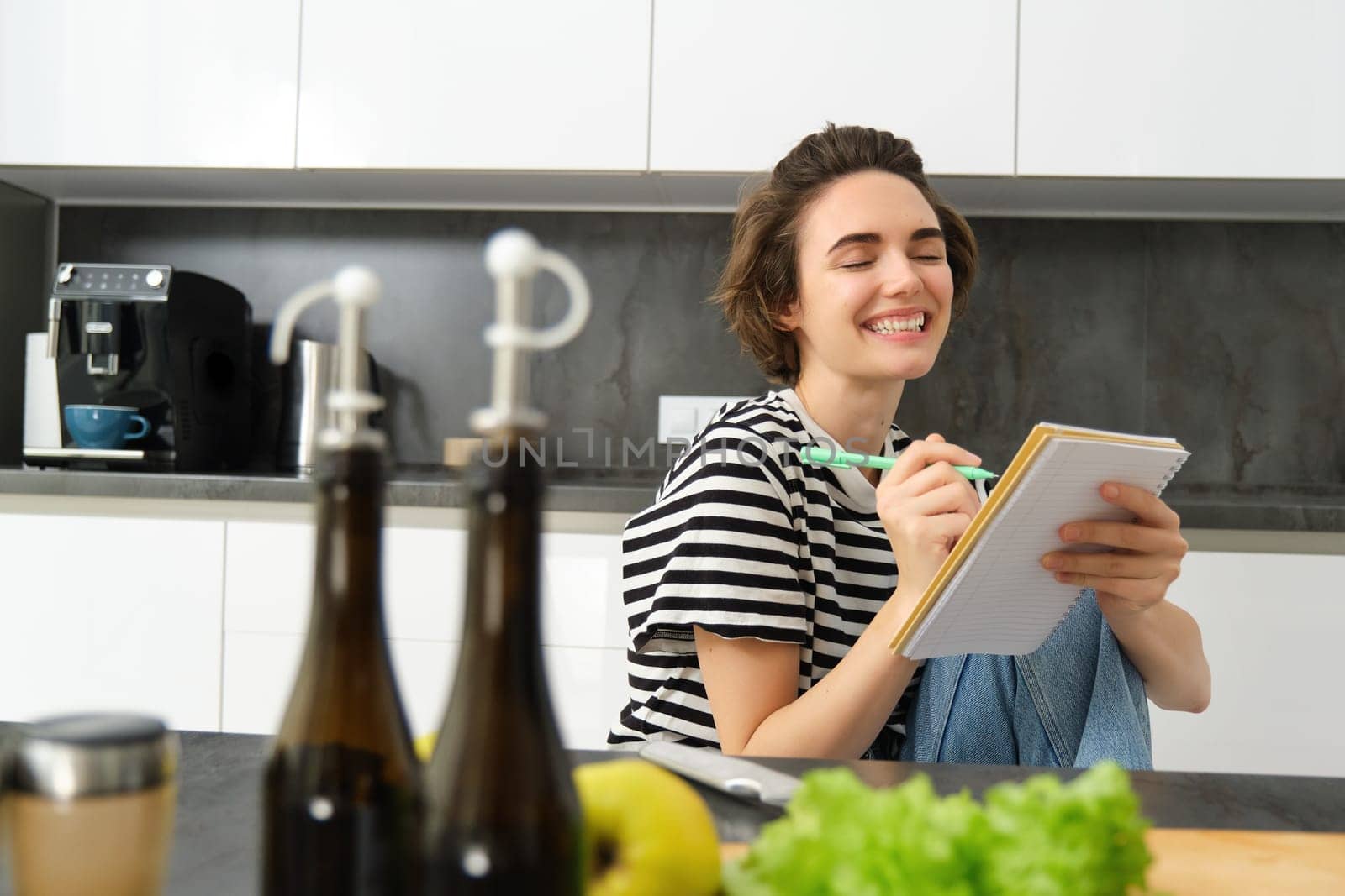 Close up of happy, smiling young woman writing in notebook, creating recipe, list of meals for this week, sitting near vegetables and olive oil, cooking in the kitchen by Benzoix