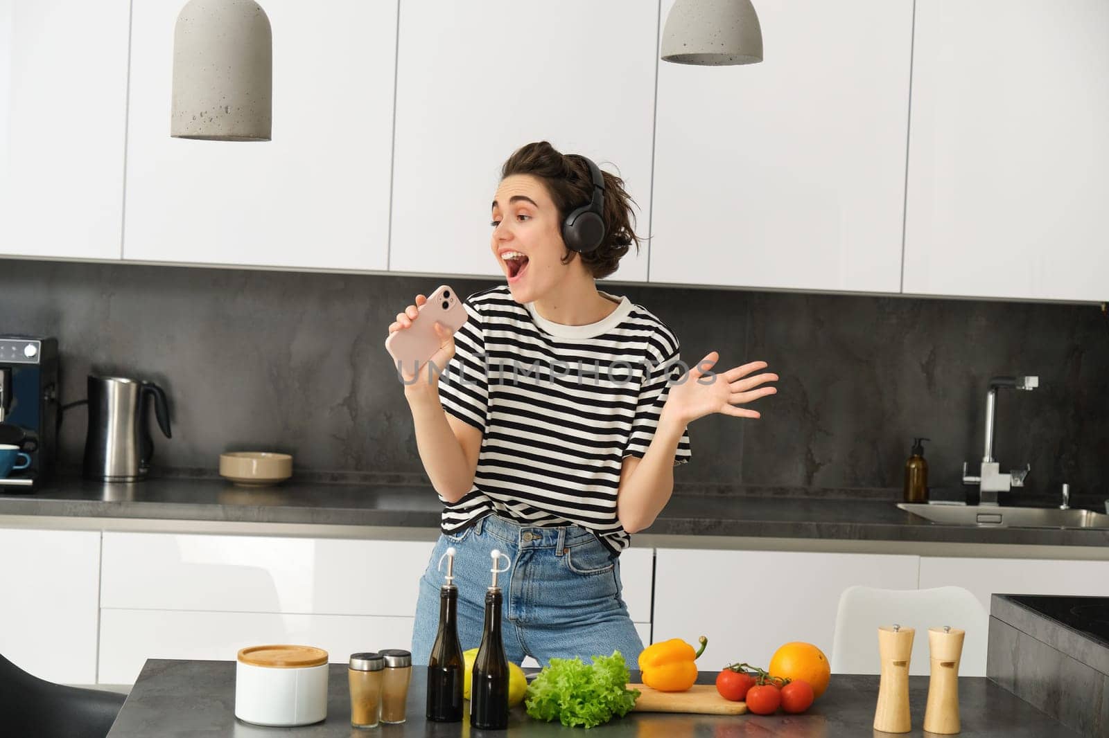 Portrait of woman singing and dancing while cooking food in the kitchen, holding smartphone, chopping vegetables on kitchen counter by Benzoix