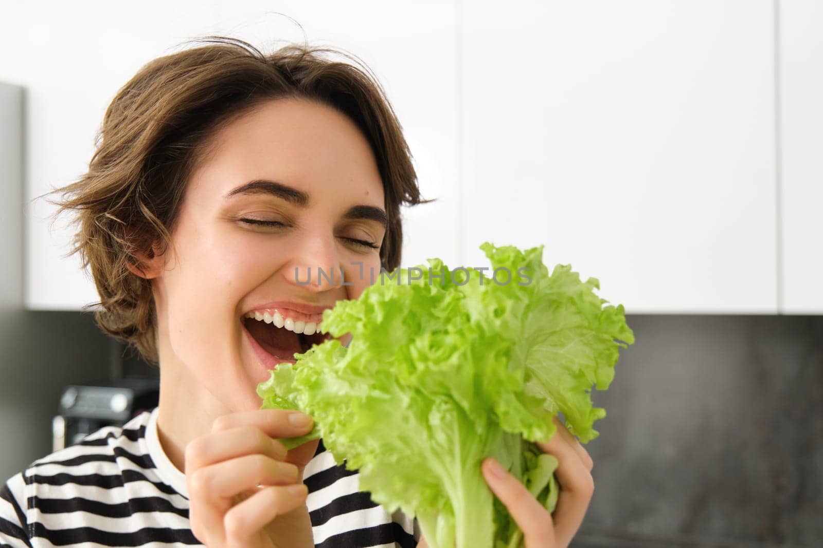 Close up portrait of happy, smiling young woman biting salad leaf, loves vegetables, making herself a salad, standing in the kitchen, cooking vegetarian meal.
