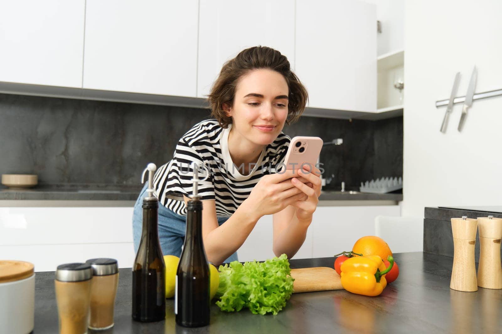 Woman bending on kitchen counter with smartphone, looking for healthy recipe online on mobile phone, standing next to chopping board and vegetables, cooking ingredients by Benzoix