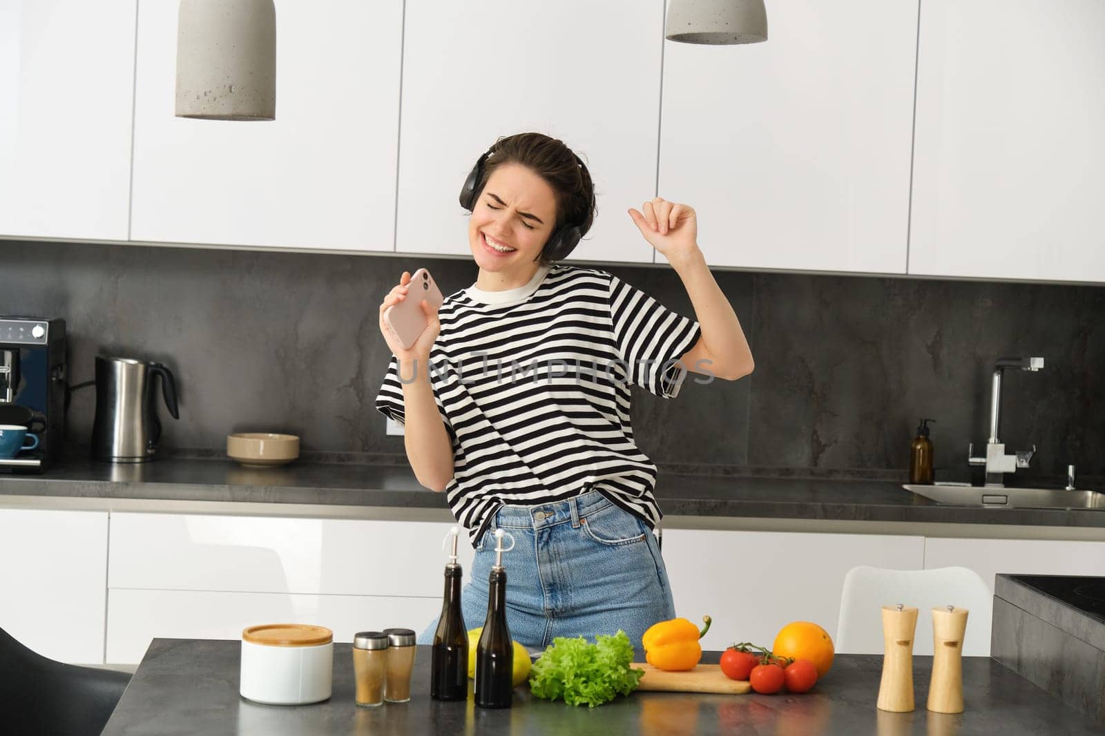 Carefree modern woman in wireless headphones, listens to music, dancing and cooking salad, preparing food in the kitchen, singing in smartphone.