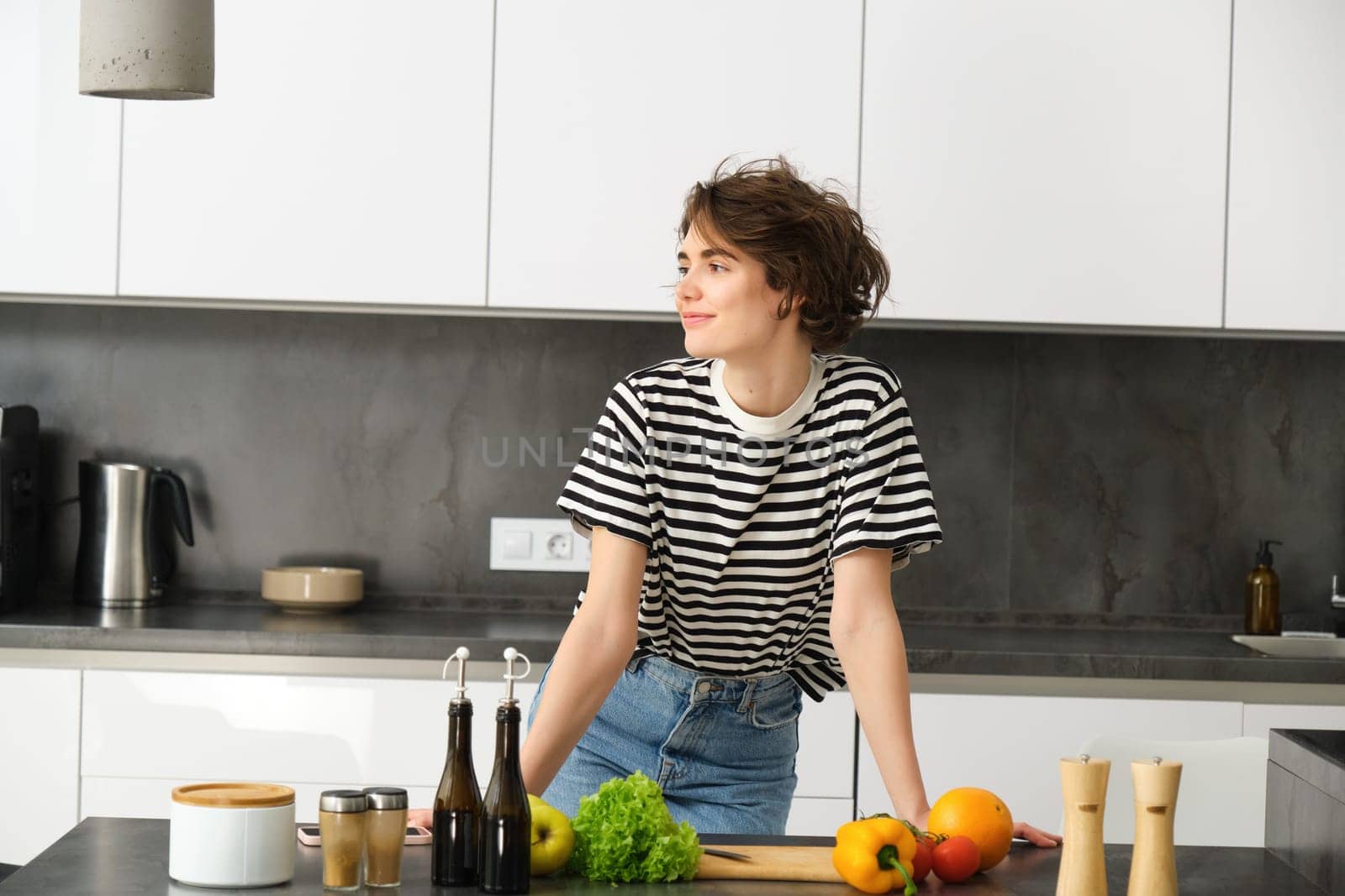 Portrait of young modern woman in the kitchen, leaning on counter with chopping board, making a salad with vegetables and dressings, cooking vegetarian meal.