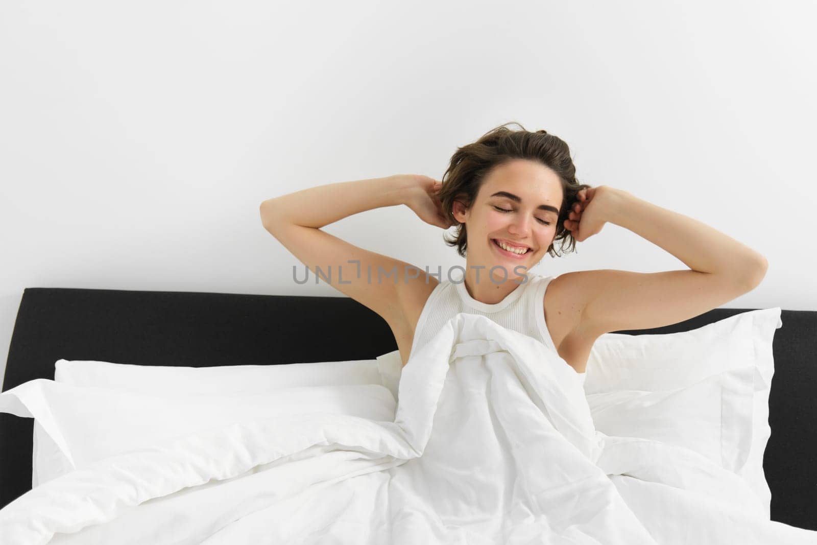 Portrait of woman feeling satisfied after good sleep, stretching hands after waking up, sitting on bed under white linen blanket, relaxing at home.