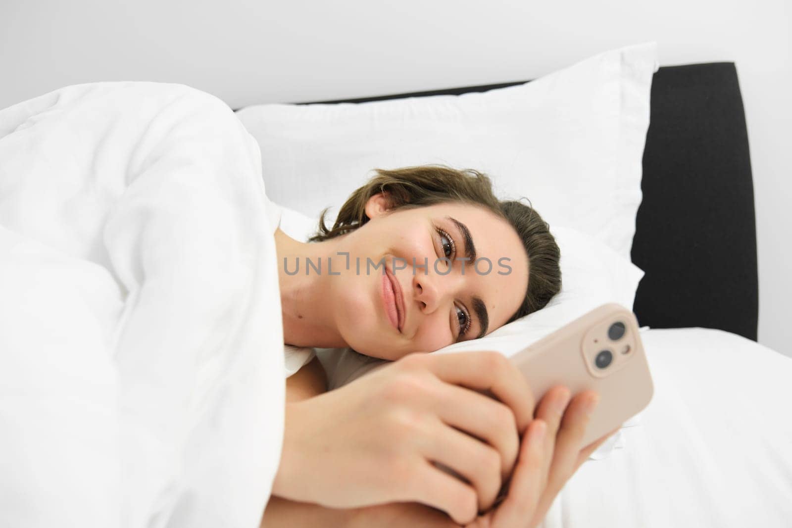 Close up portrait of lovely young woman in bed, lying and going to sleep, checking her phone messages, using smartphone and smiling by Benzoix