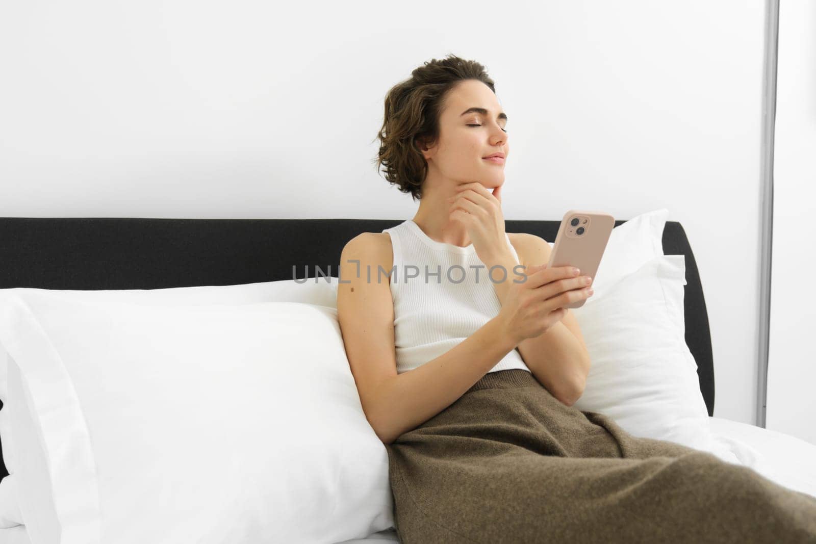 Portrait of young woman with eyes closed and pleased smile, holding smartphone, lying and resting in bed, using mobile phone.