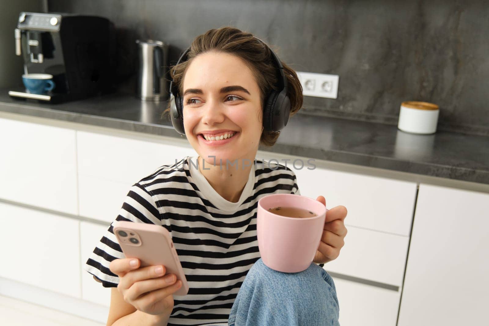 Stylish young woman in headphones, listens music and uses smartphone, drinks warm tea in kitchen, relaxing at home.