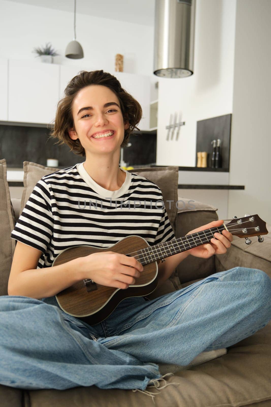 Vertical shot of young musician, woman learns how to play ukulele, sits on sofa with crossed legs and smiling at camera.