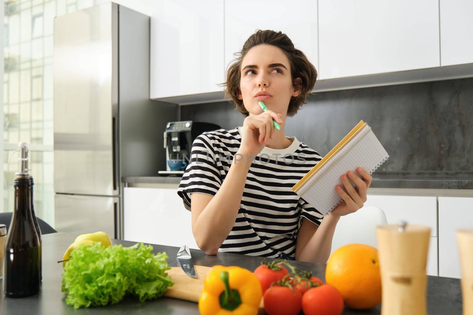 Portrait of thinking woman with notebook, cooking, writing down recipe ingredients, deciding on a meal for dinner, sitting near vegetables and chopping board in kitchen by Benzoix