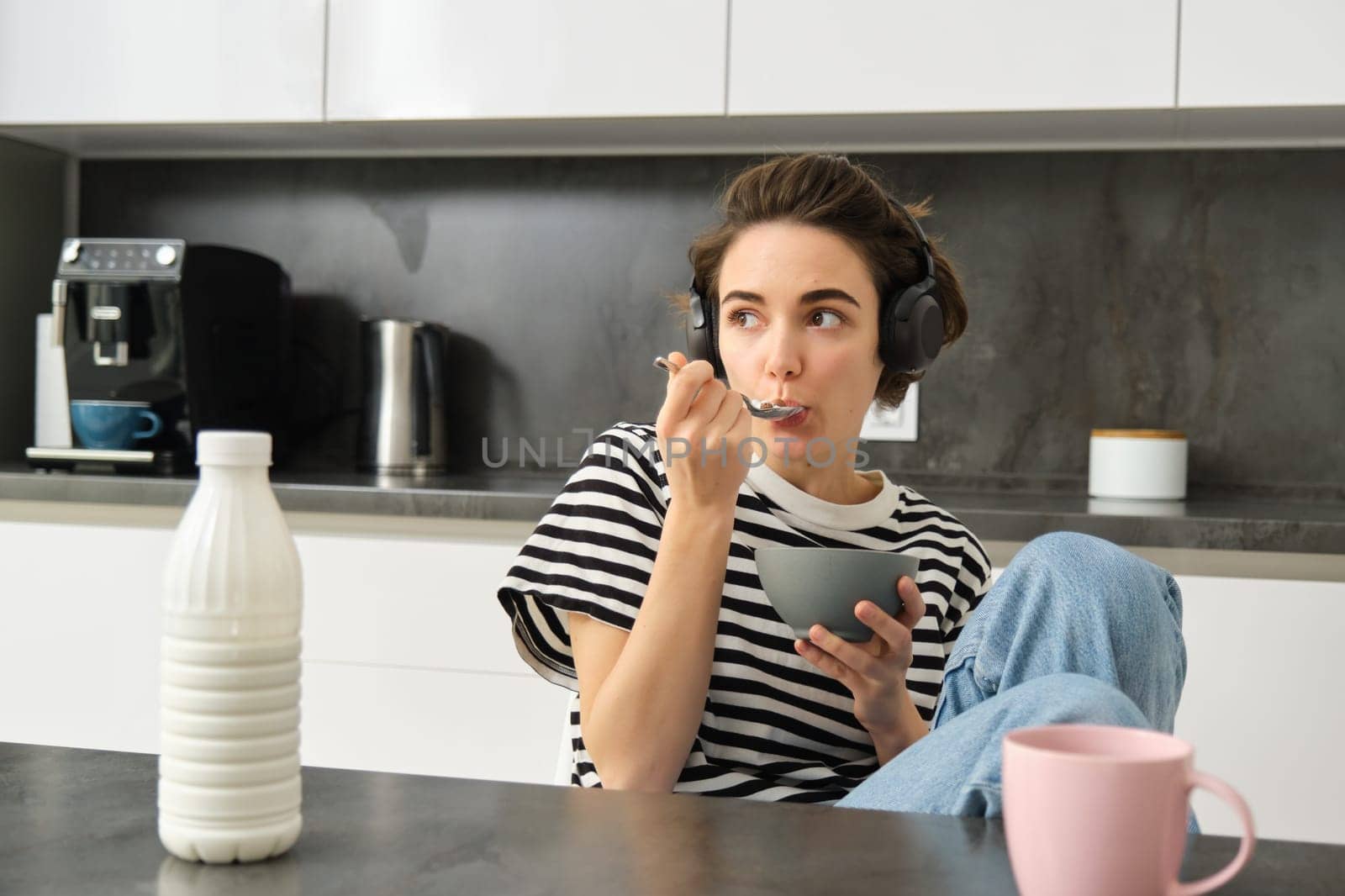 Cute modern woman, student eating quick breakfast, having cereals with milk and coffee, listening to podcast or music in wireless headphones.