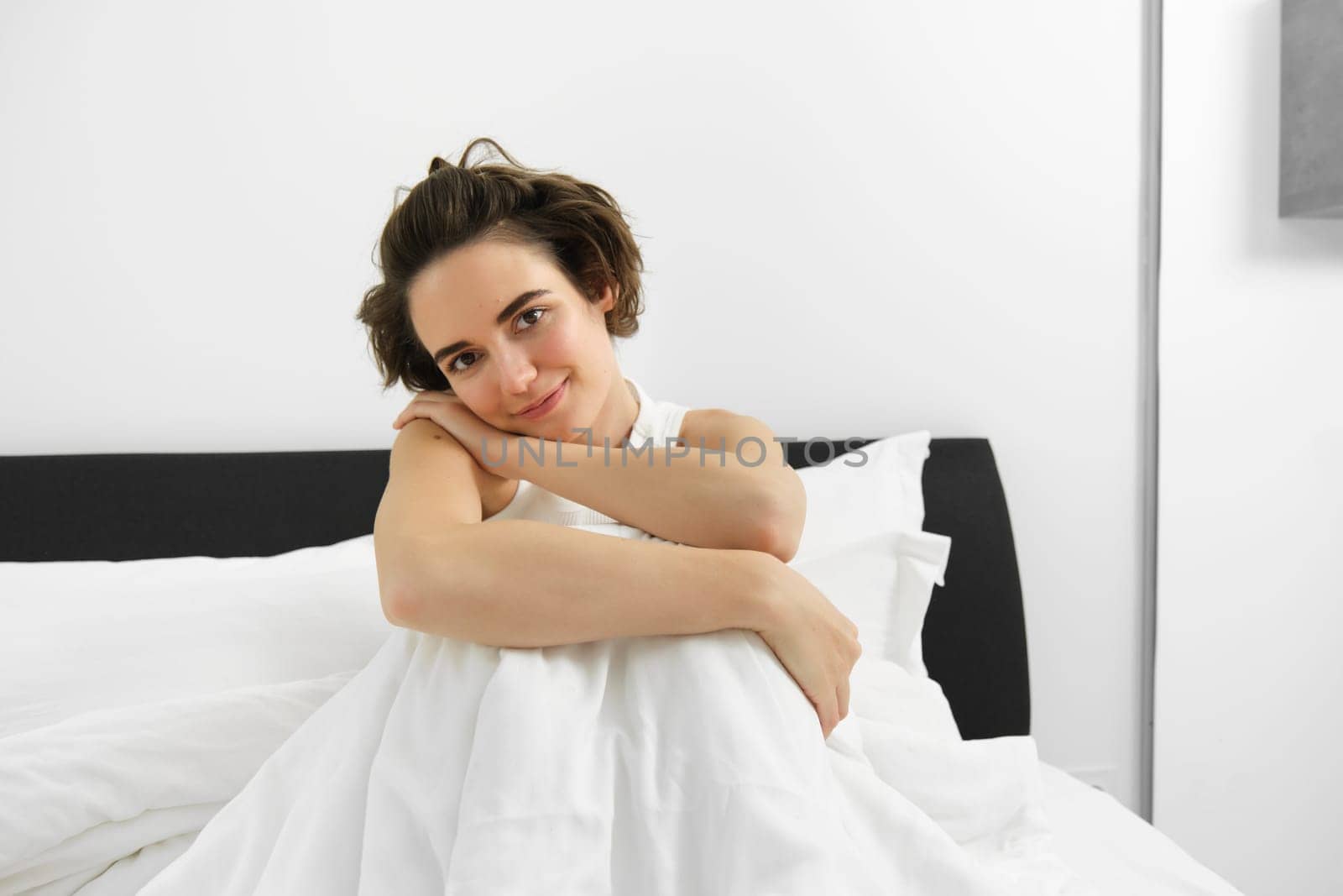 Image of beautiful brunette woman sitting on bed, covered in white linen sheets, looking at camera with dreamy, sensual smile, gazing at someone. Morning and lifestyle concept