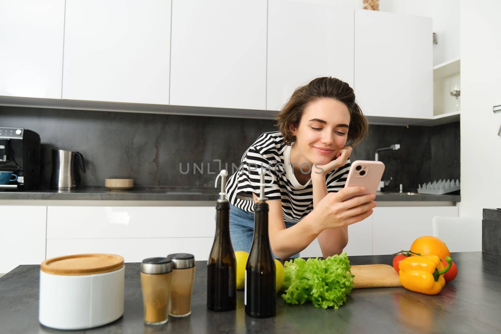 Girl cooking healthy meal in the kitchen, reading recipe on smartphone, using mobile phone and making meal from fresh green vegetables.