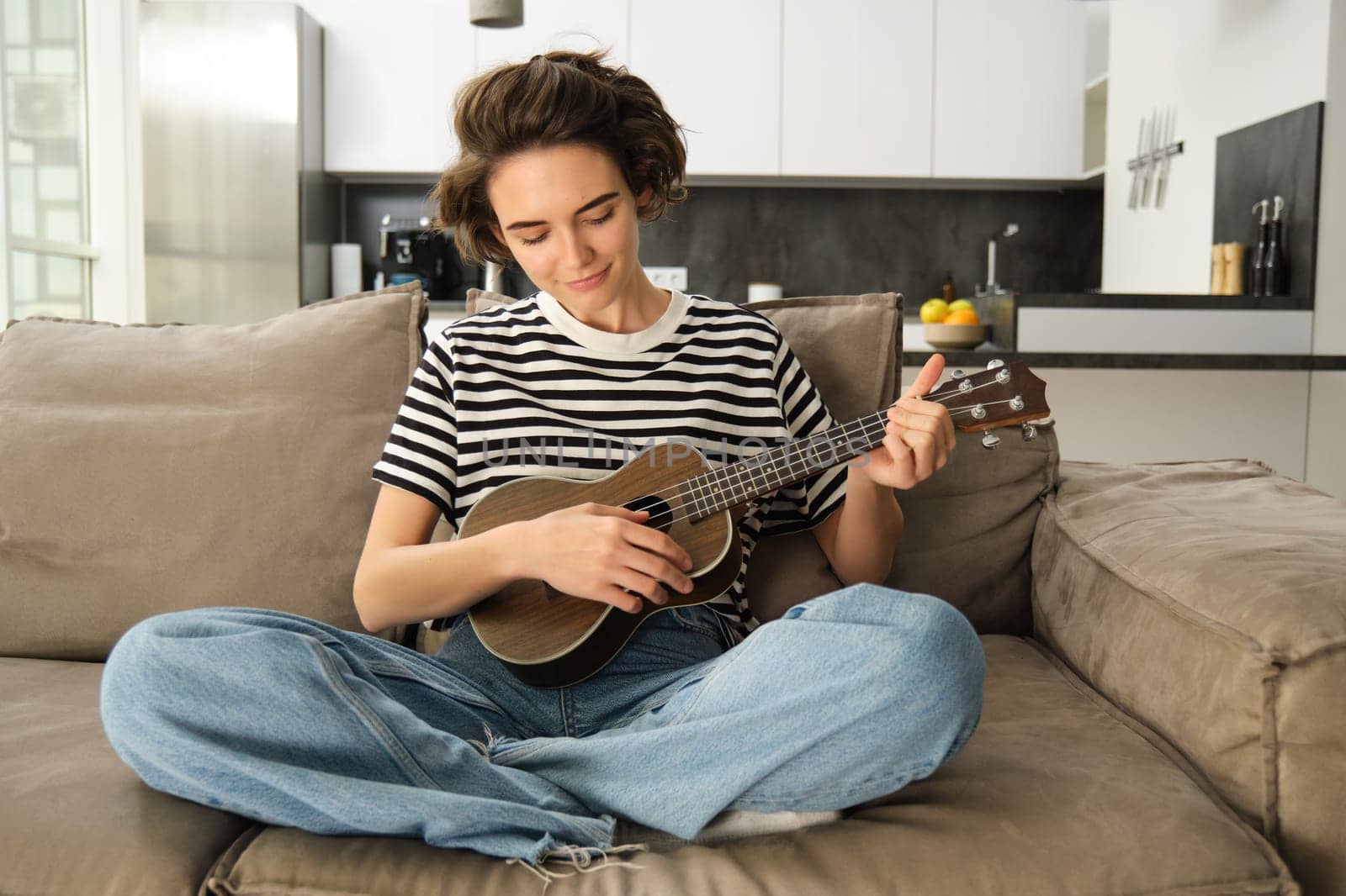 Lifestyle and hobbies concept. Young smiling woman on sofa, playing ukulele, singing and learning strumming pattern for favourite song by Benzoix