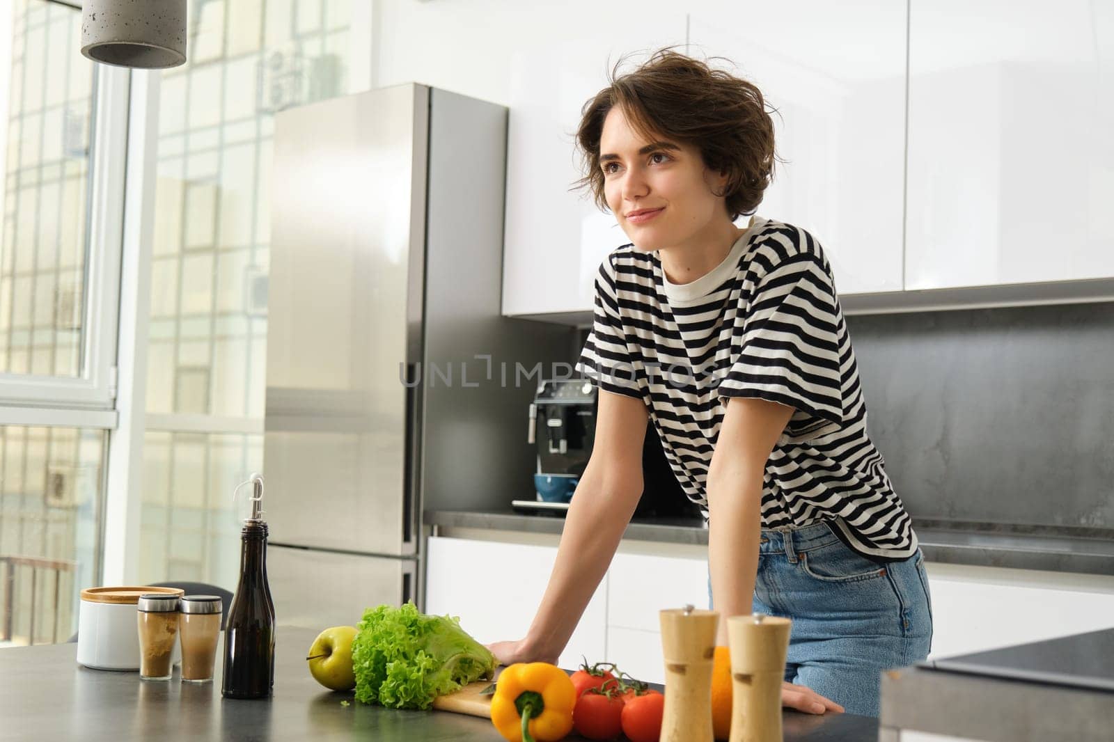 Portrait of beautiful young woman, female model posing near chopping board with vegetables and salad dressing, making herself light diet meal, vegan food for breakfast, standing in kitchen by Benzoix