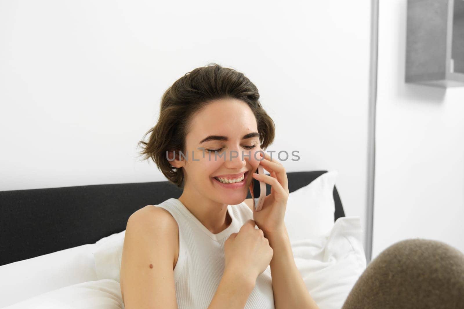 Female model sitting on bed with phone, calling friend, chatting over telephone, laughing and smiling.