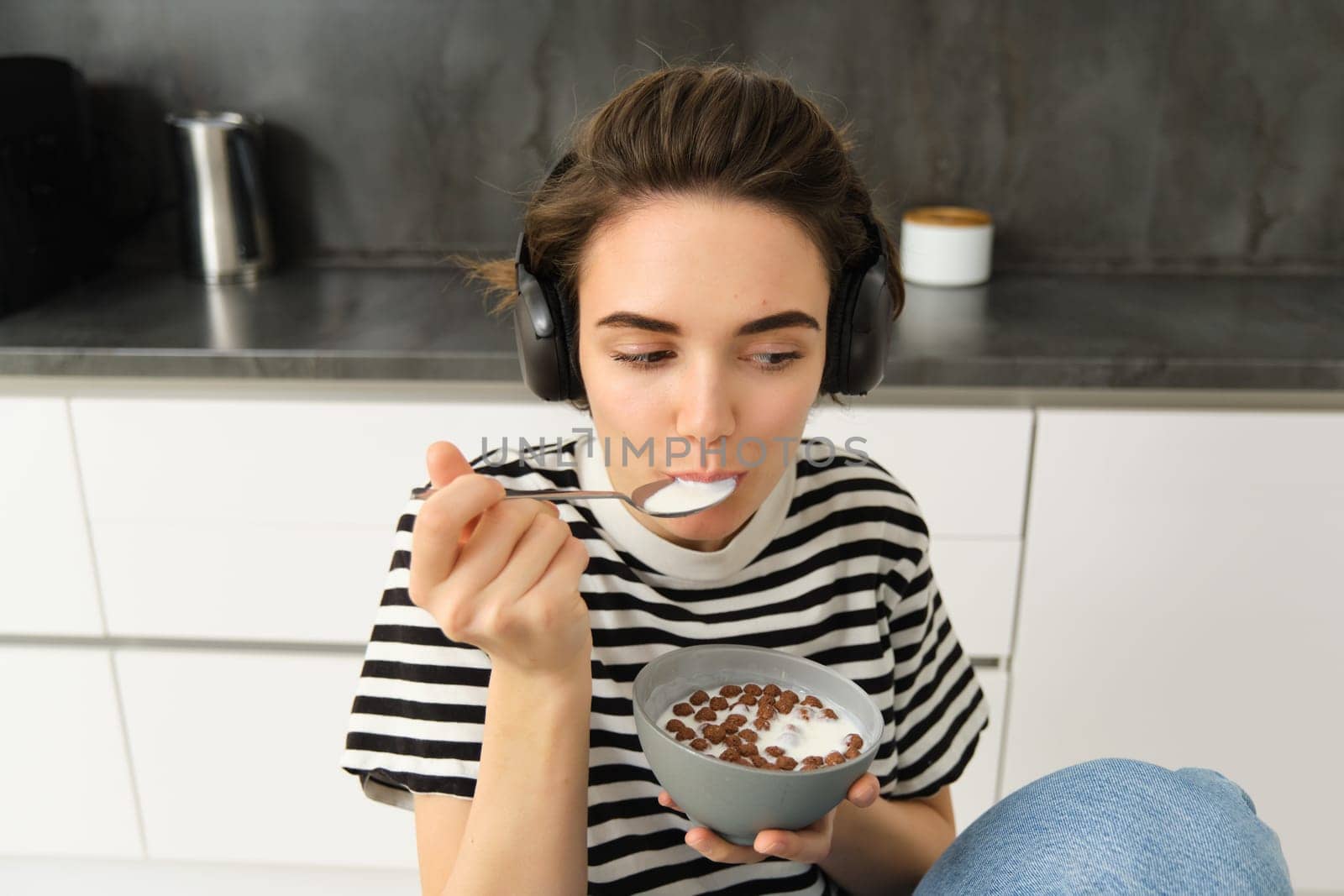 Cute brunette girl, student eats her cereals with milk for breakfast, holding spoon near mouth, listens to music in headphones, sitting on chair in kitchen.
