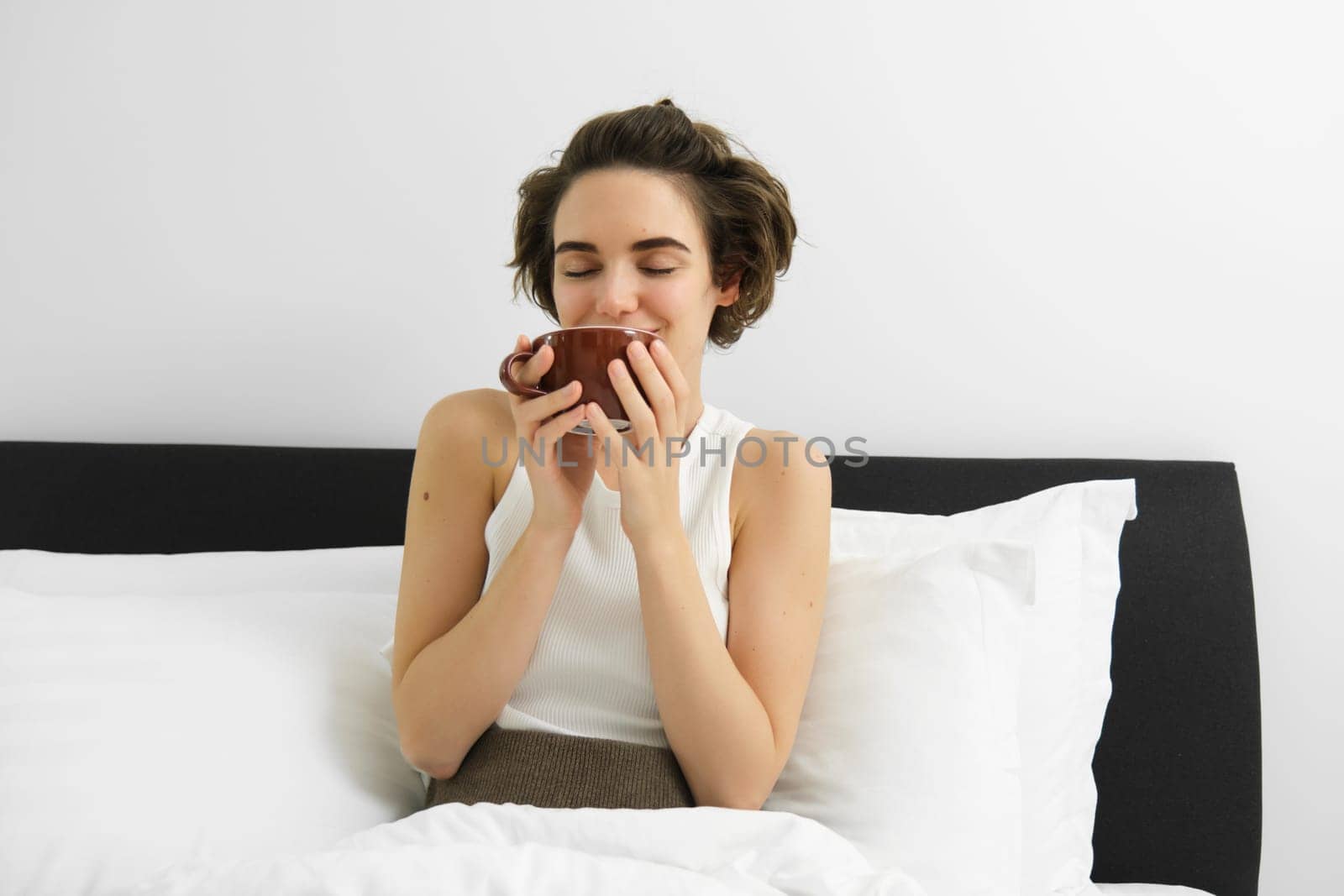 Beautiful young woman waking up and drinking coffee in her bed, smiling from satisfaction. by Benzoix