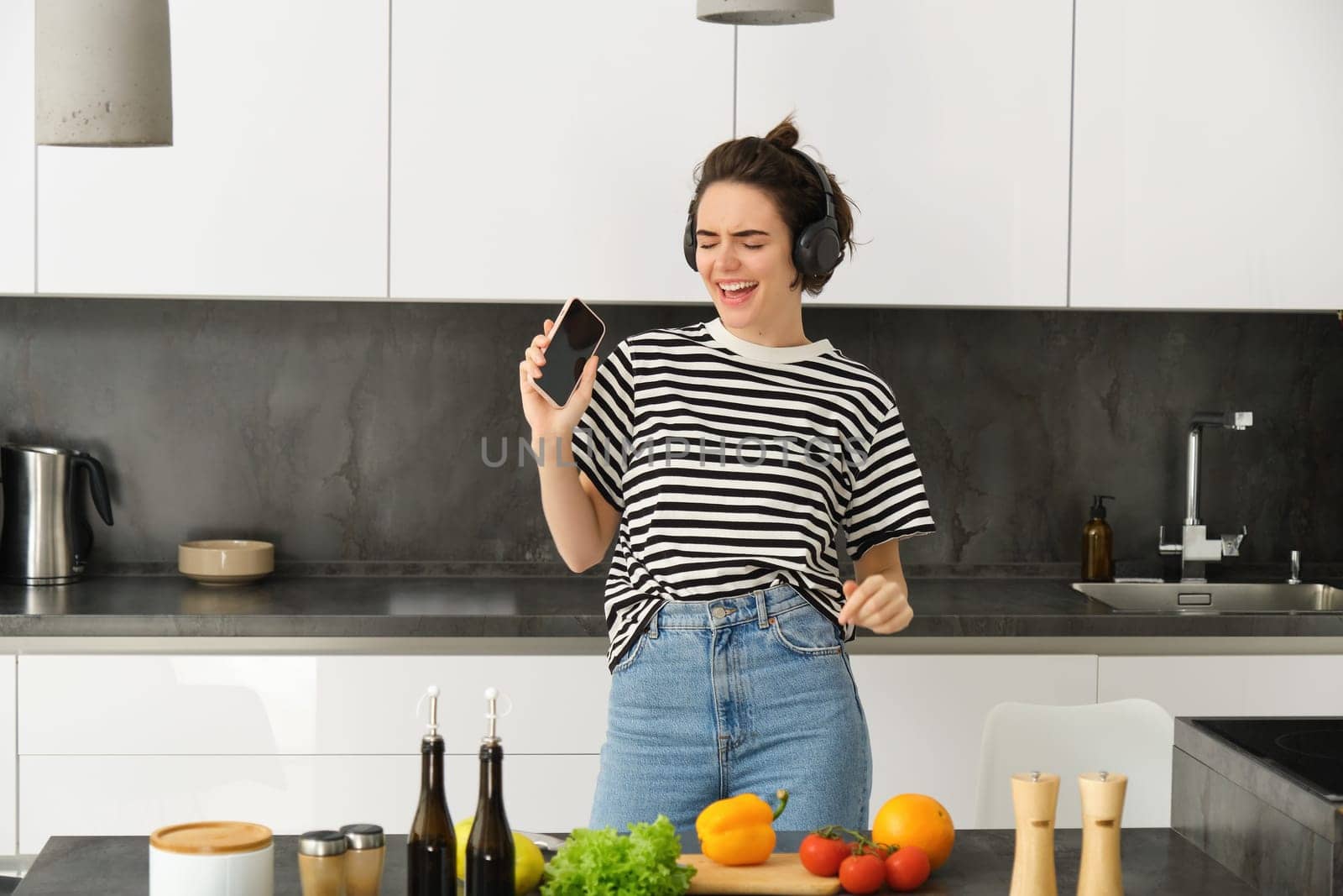 Portrait of happy girl cooking salad and listening to music in headphones, dancing while making a meal in the kitchen.