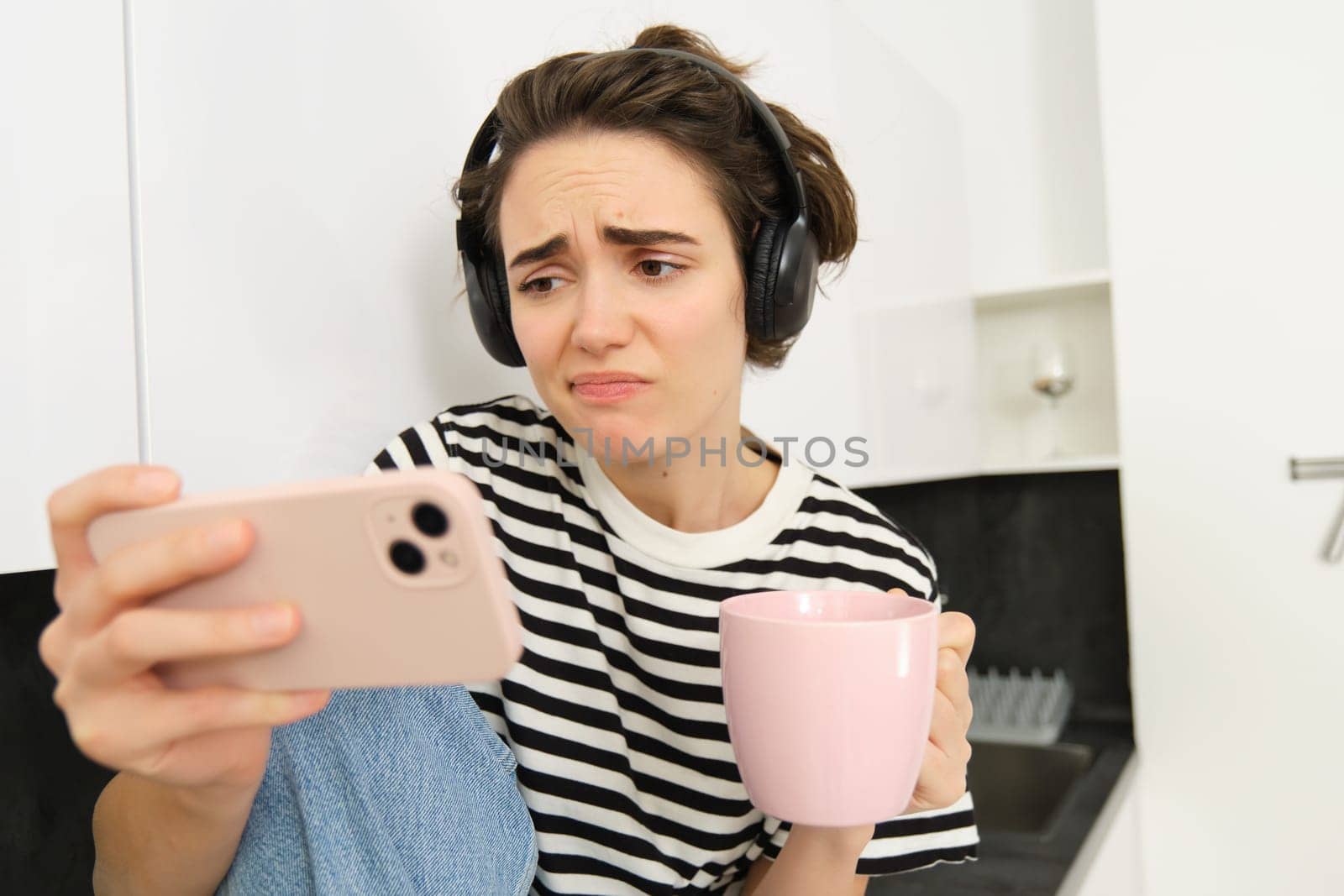 Portrait of woman looking with sympathy and sad face at smartphone screen, watching video on mobile phone app, drinking tea or coffee in the kitchen, frowning and looking with pity.