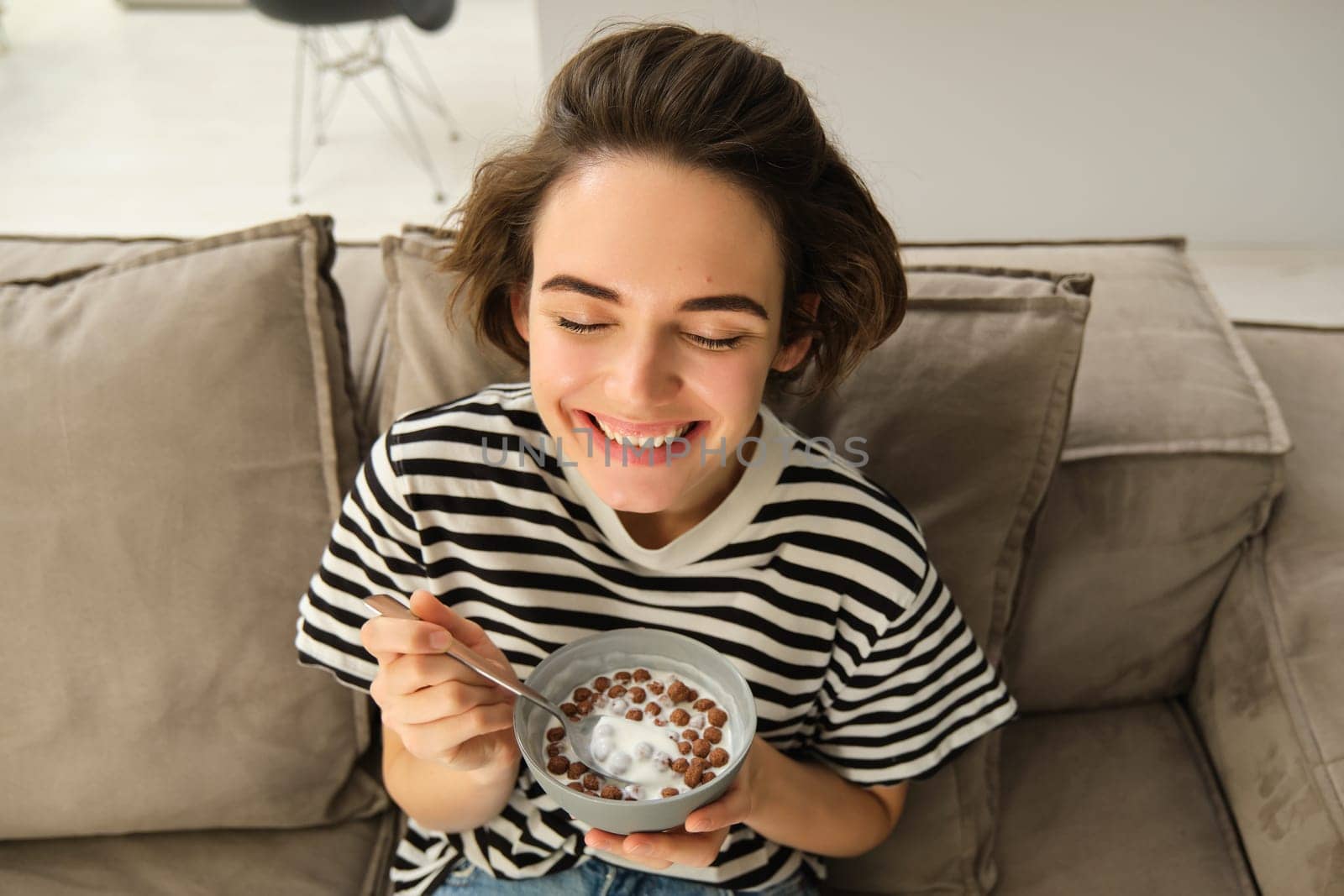 Upper angle shot of happy, cute young woman on sofa, eating bowl of cereals with milk and smiling, enjoying her breakfast by Benzoix