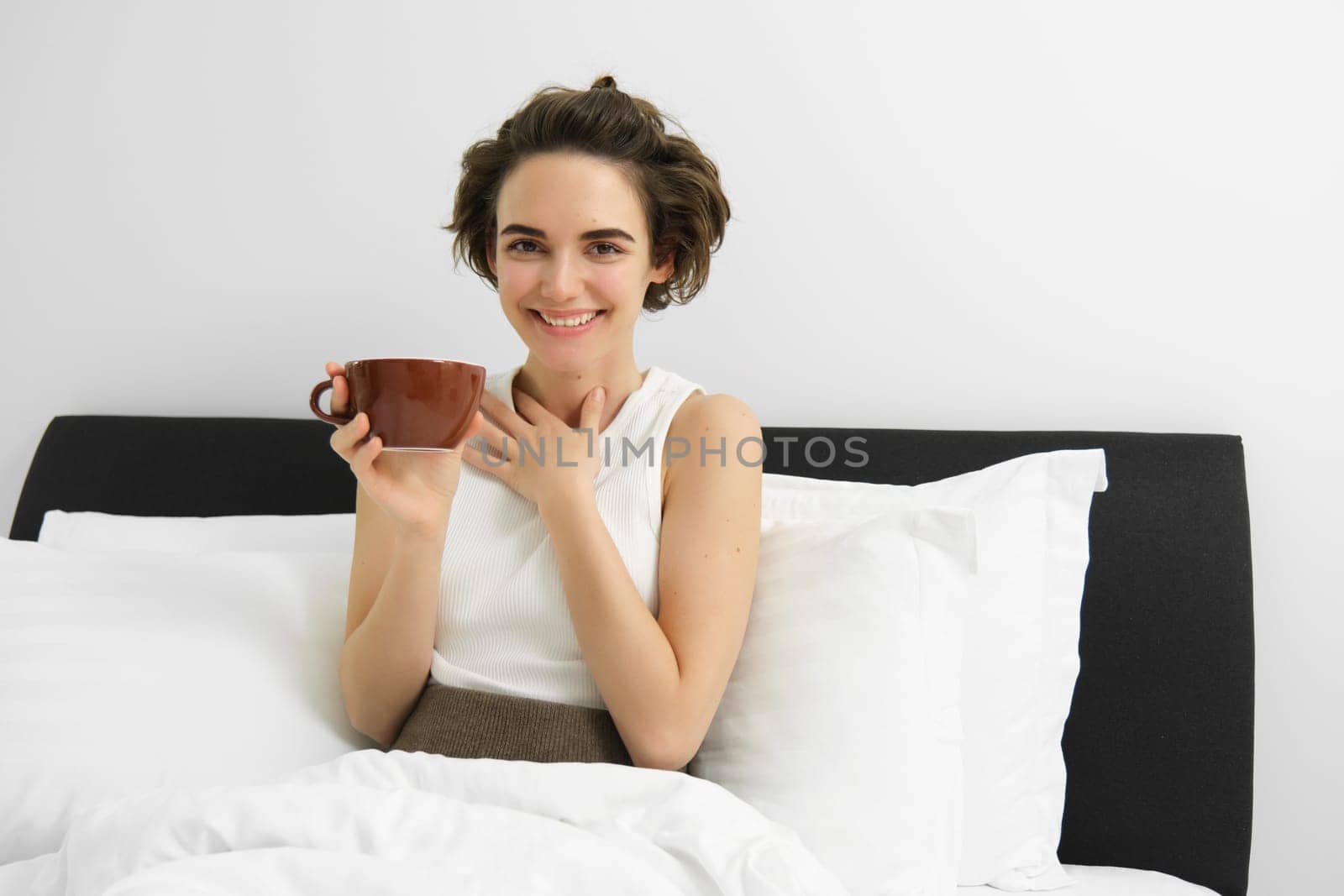 Beautiful young woman with cup of coffee, drinking cappuccino in bed and smiling.