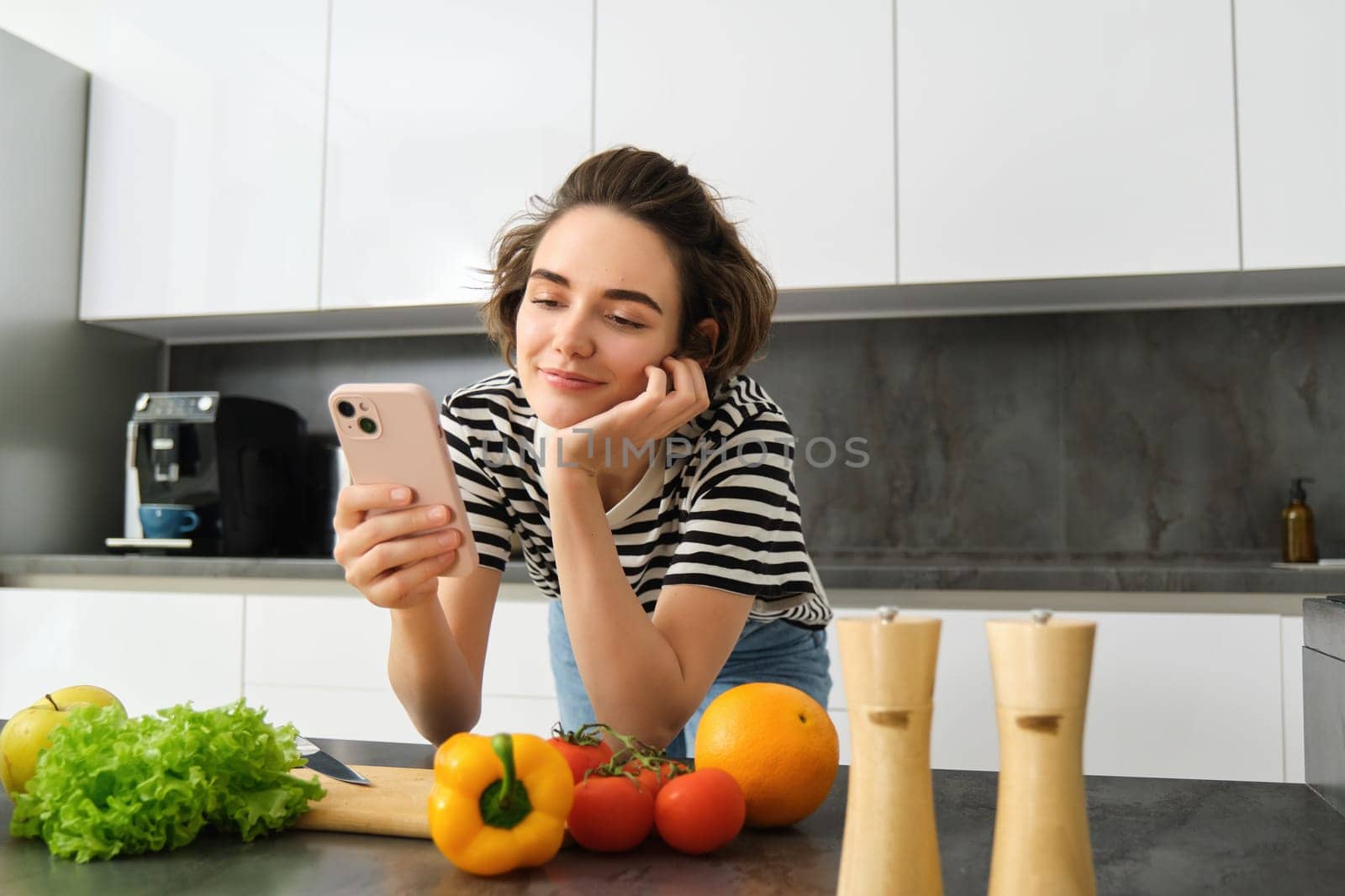 Portrait of beautiful young woman with smartphone, cooking in the kitchen, chopping vegetables, reading recipe online.