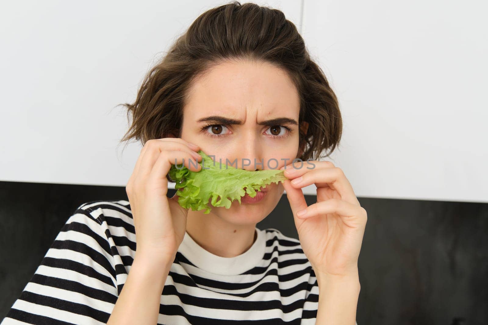 Frowning silly woman, holding lettuce leaf near face and frowning at camera, making pouting face, dislike eating vegetables.