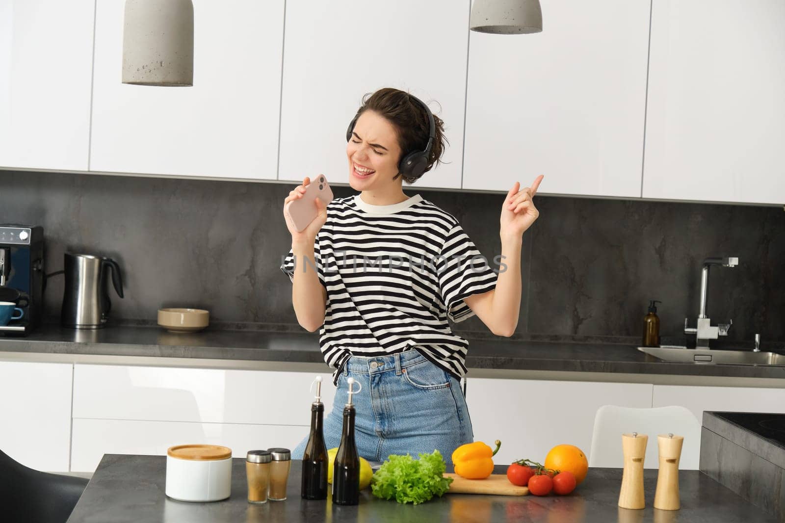 Portrait of happy girl cooking salad and listening to music in headphones, dancing while making a meal in the kitchen by Benzoix