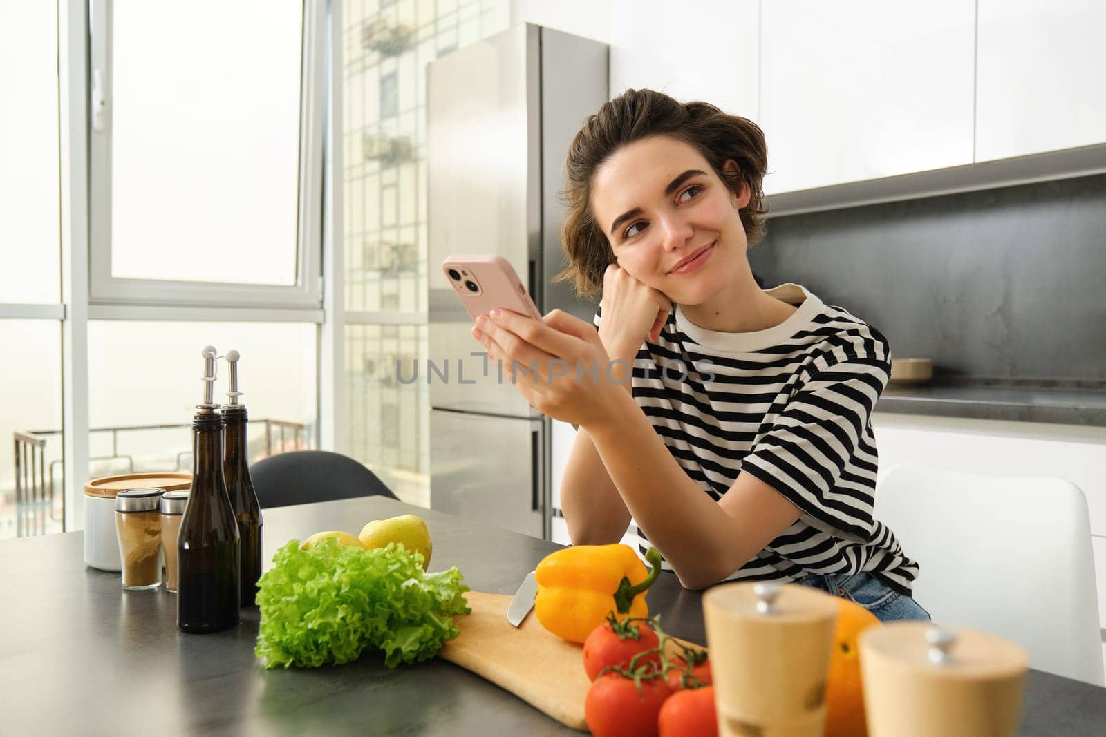 Portrait of brunette girl cooking food in the kitchen, searching recipes on social media app, holding mobile phone, standing near chopping board and vegetables, preparing healthy vegetarian meal.