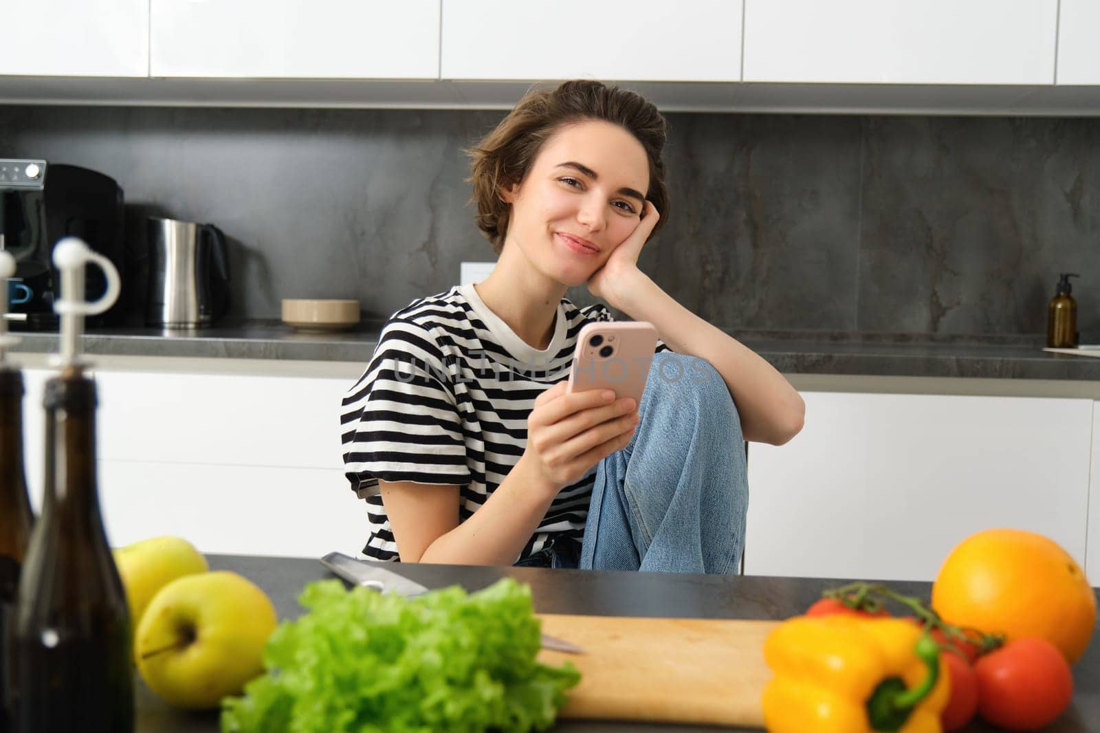 Portrait of young woman searching for cooking recipes online using smartphone, sitting near vegetables, salad ingredients and chopping board, smiling at camera.