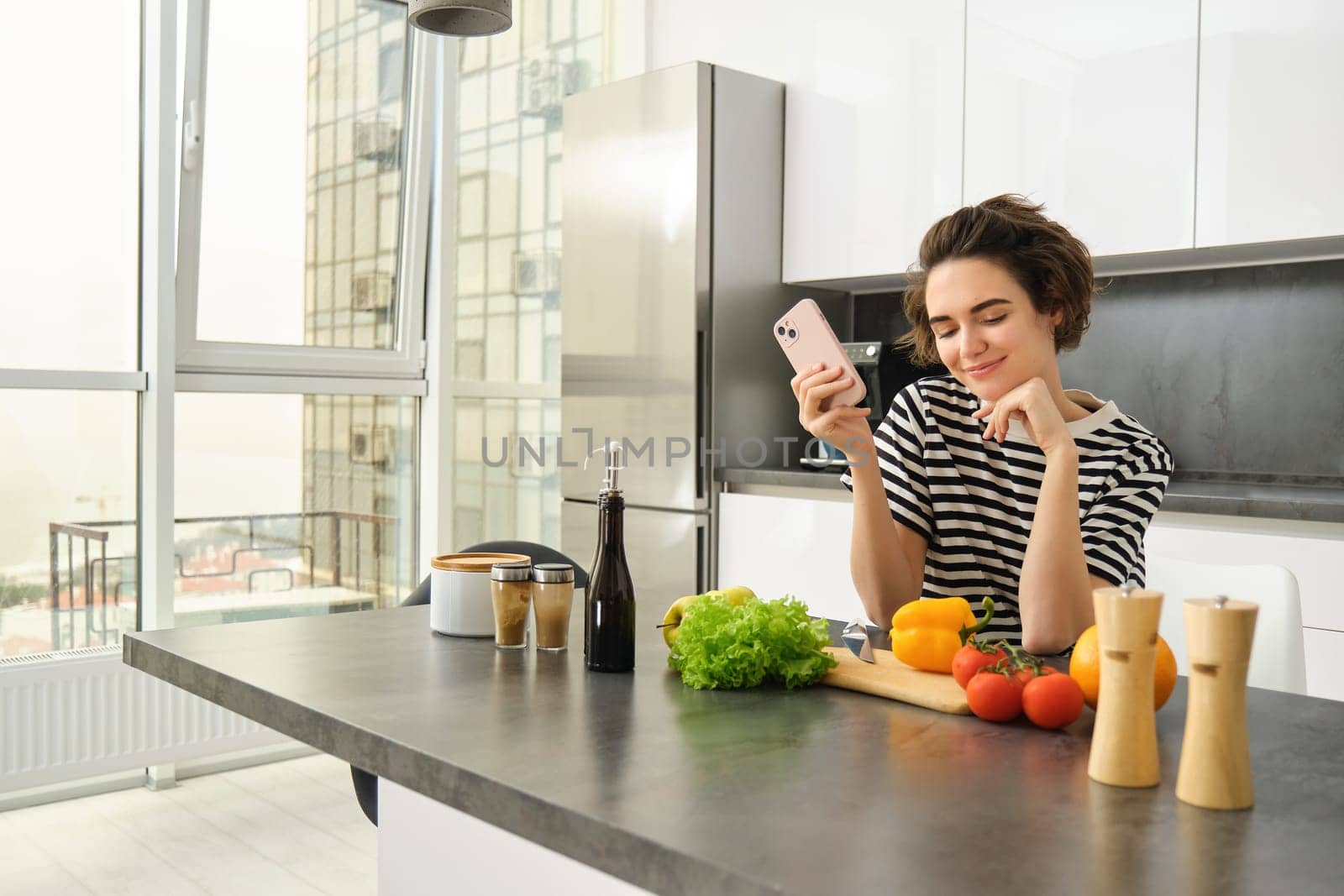 Portrait of smiling beautiful woman in the kitchen, looking at vegetables and holding smartphone, deciding what to cook, searching for ideas for lunch meals.