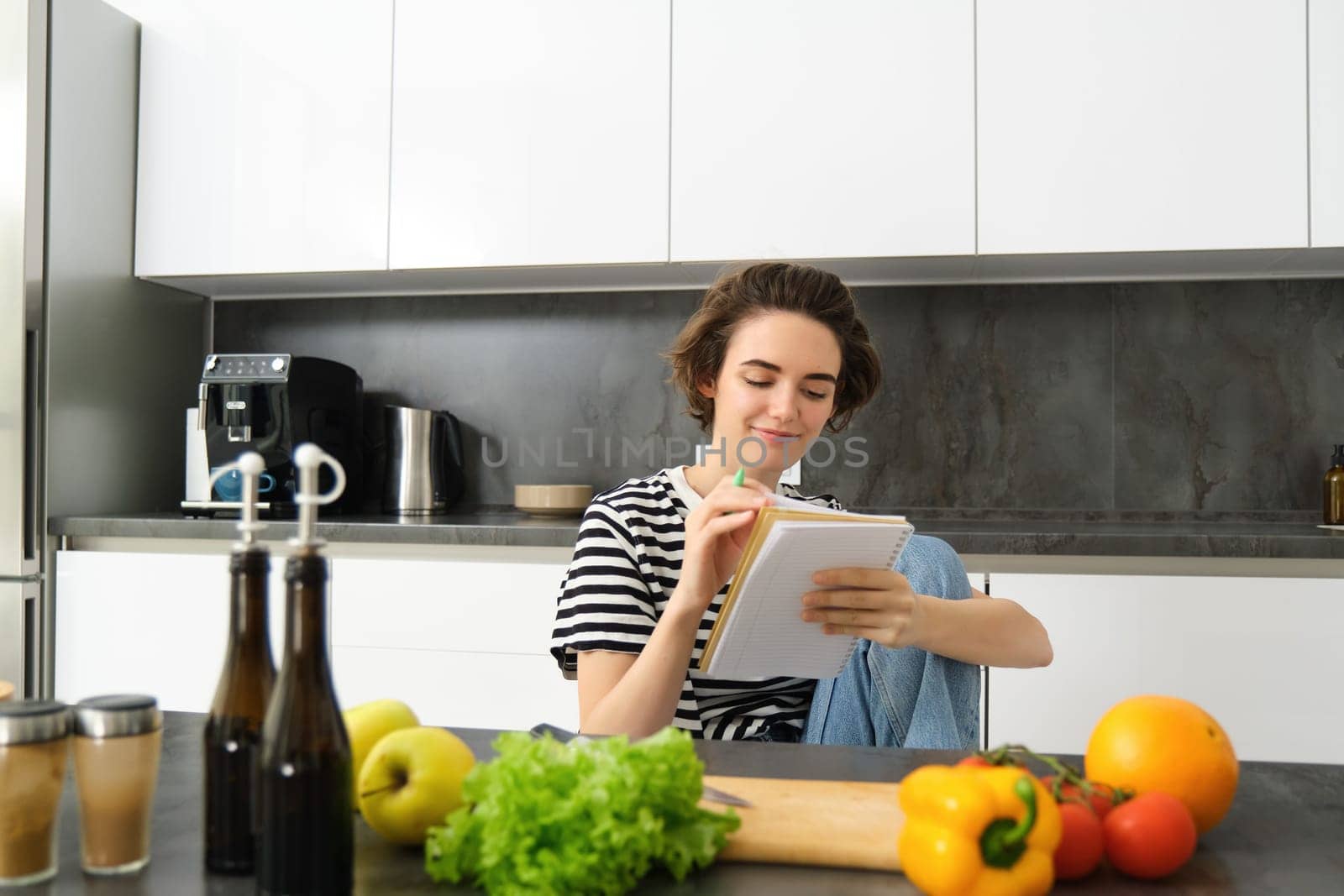 Portrait of young smiling woman in kitchen, holding notebook, making notes for recipe, writing grocery list, cooking salad, sitting near vegetables and chopping board by Benzoix