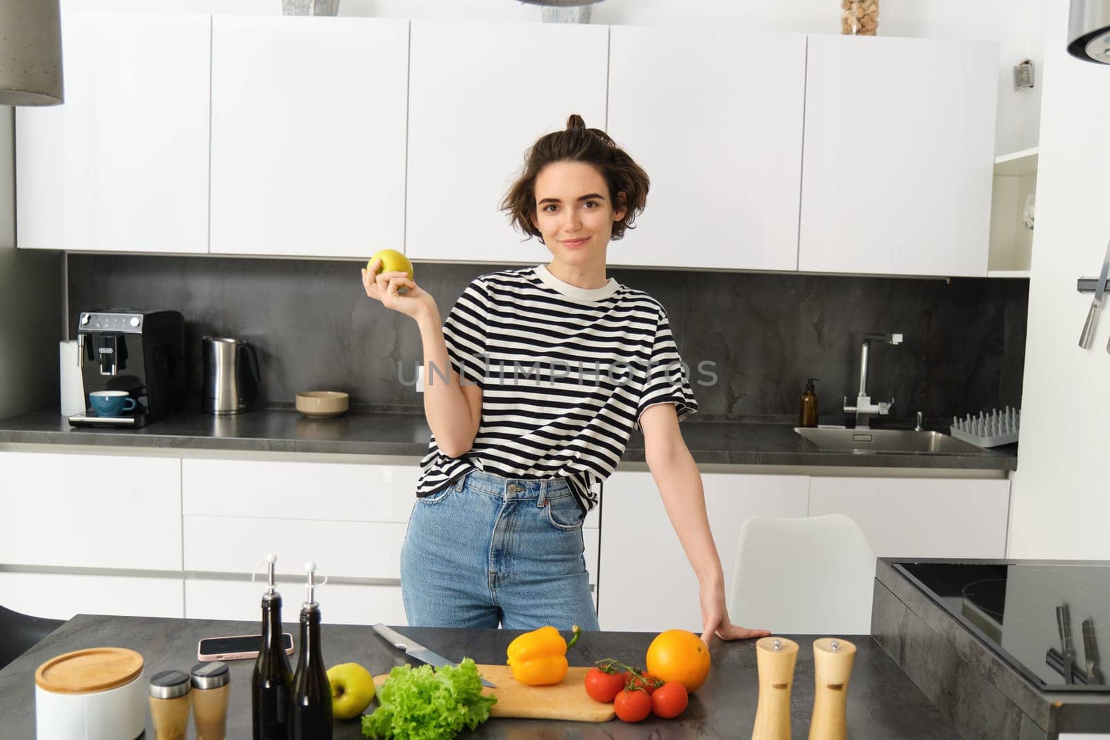 Portrait of beautiful, confident smiling woman, posing near kitchen counter with vegetables and chopping board, holding apple, cooking meal, preparing a salad.