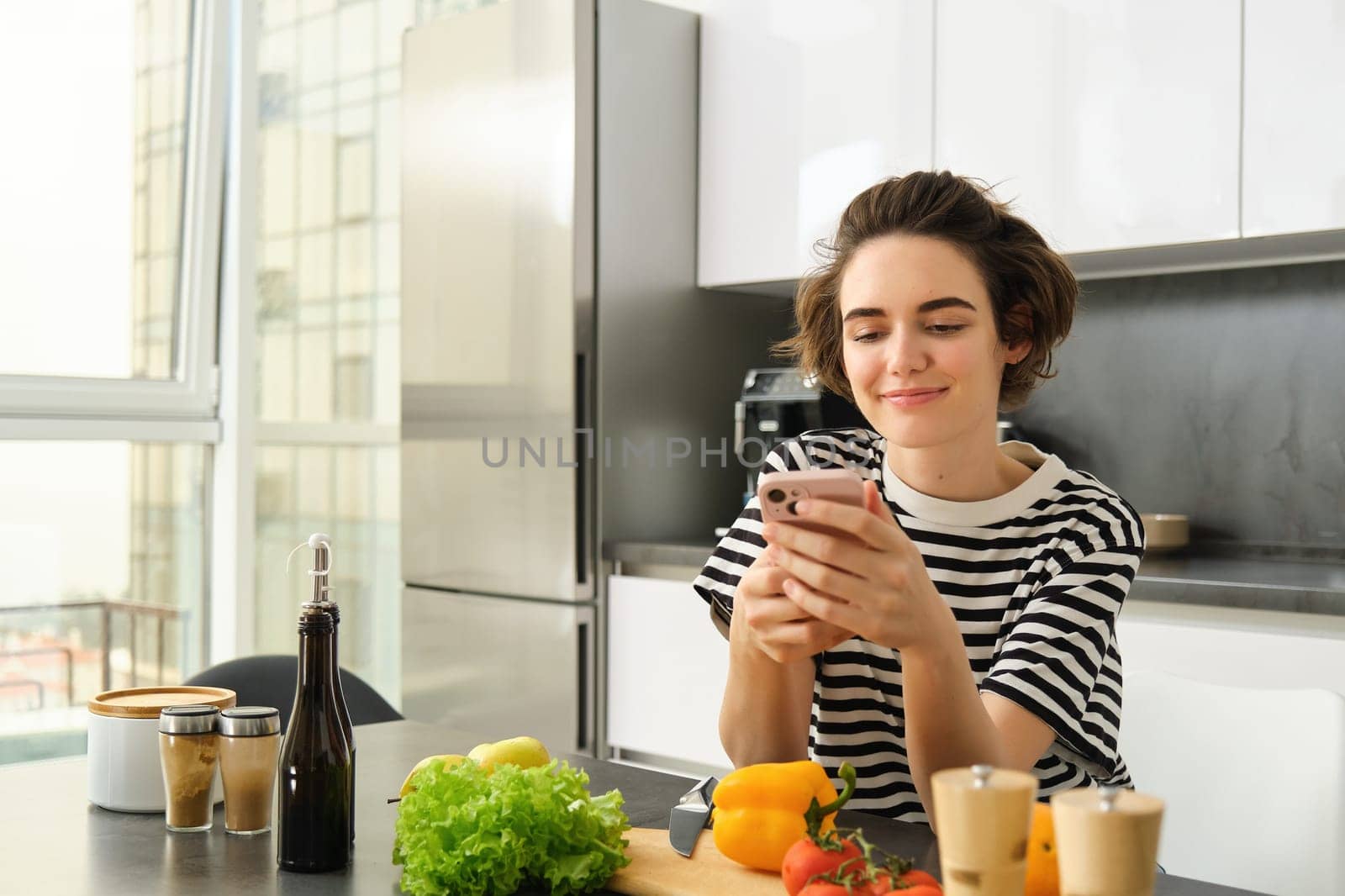 Portrait of cute modern woman cooking in the kitchen, using smartphone, reading recipe, searching meals for breakfast, leaning on counter with vegetables and chopping board.