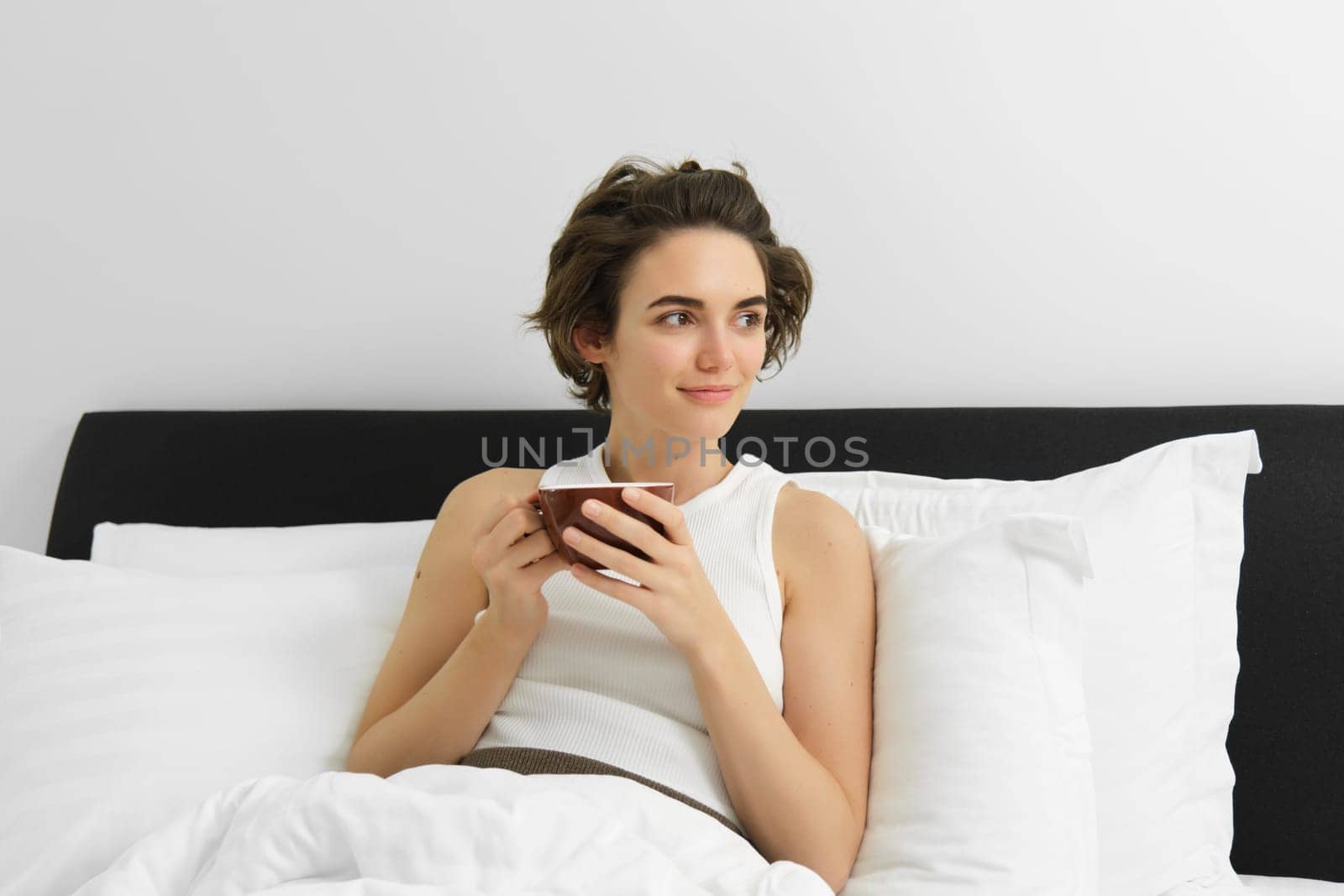 Beautiful young woman waking up and drinking coffee in her bed, smiling from satisfaction. Morning and happiness concept