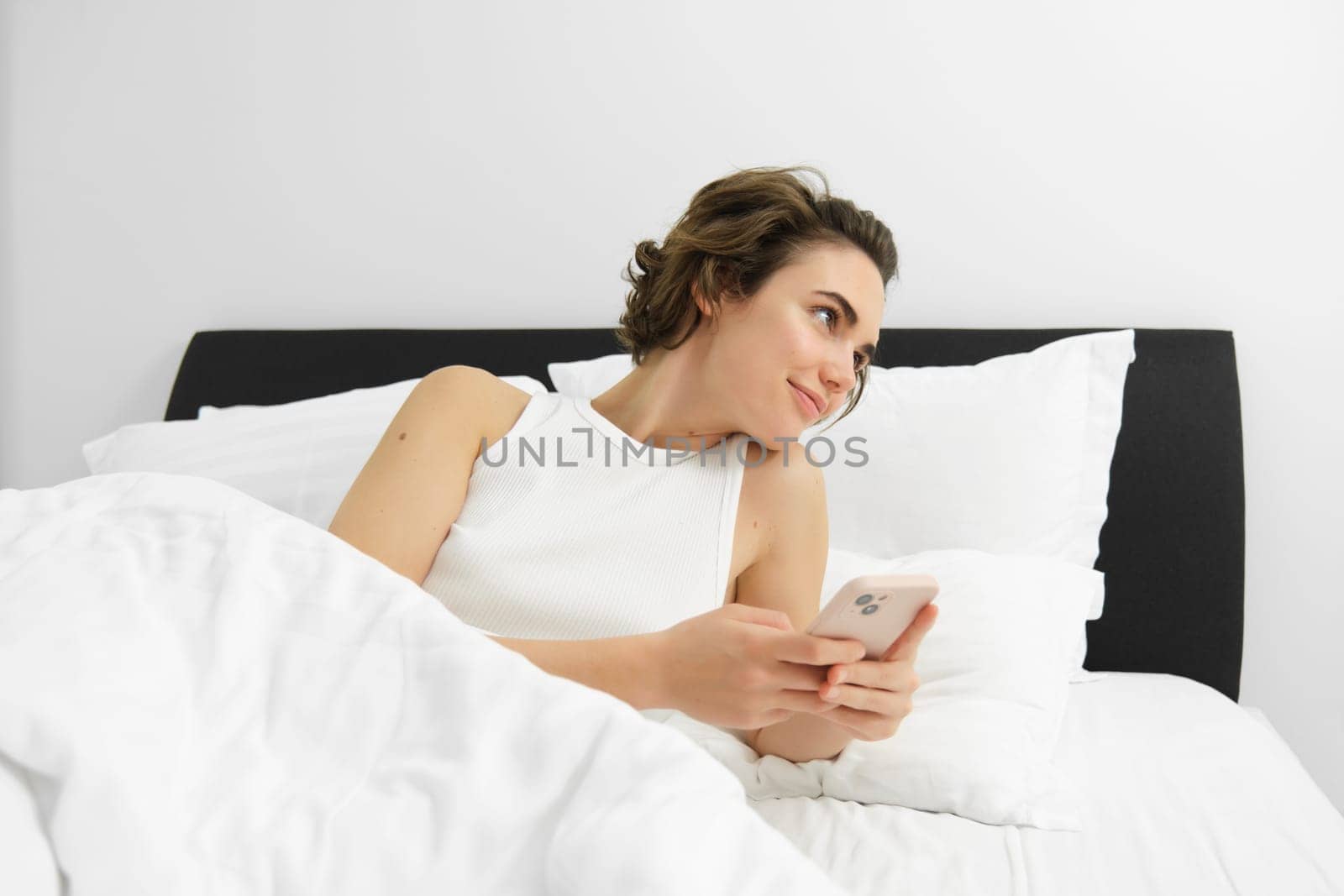 Portrait of woman in bed, lying on white sheets, using smartphone, holding mobile phone by Benzoix