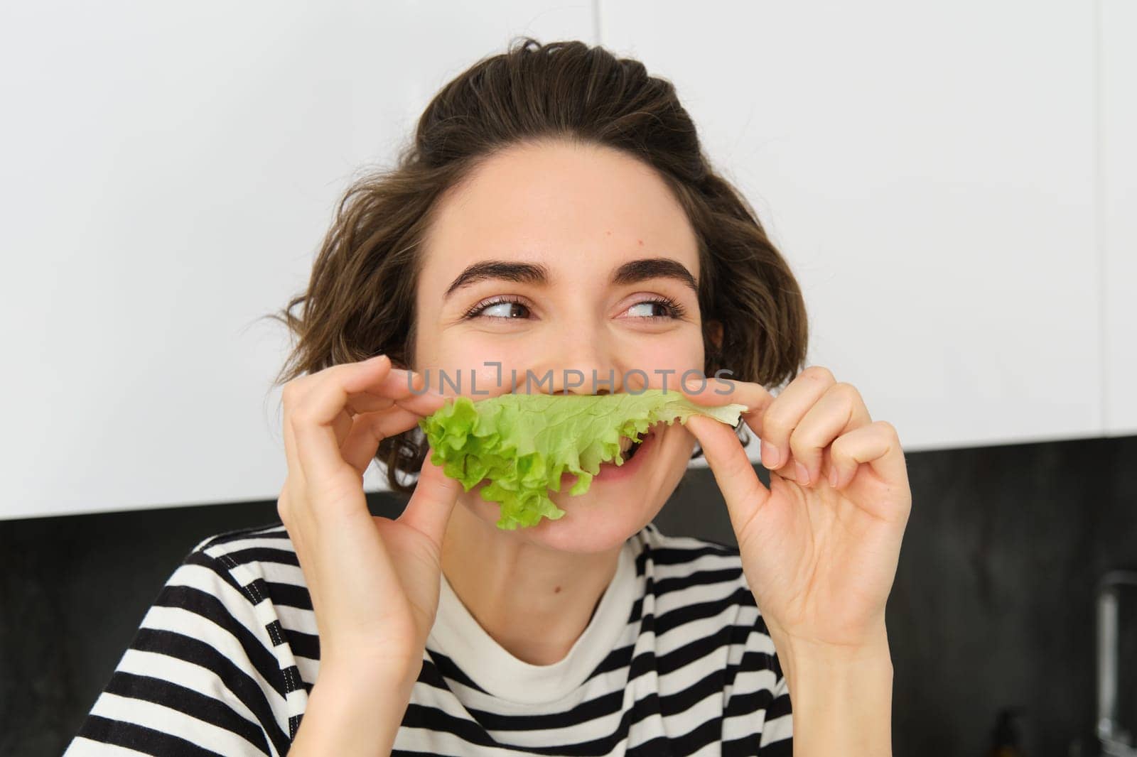 Cute smiling woman eating lettuce leaf and smiling, having a healthy snack, likes vegetables, posing in the kitchen. Lifestyle and food concept