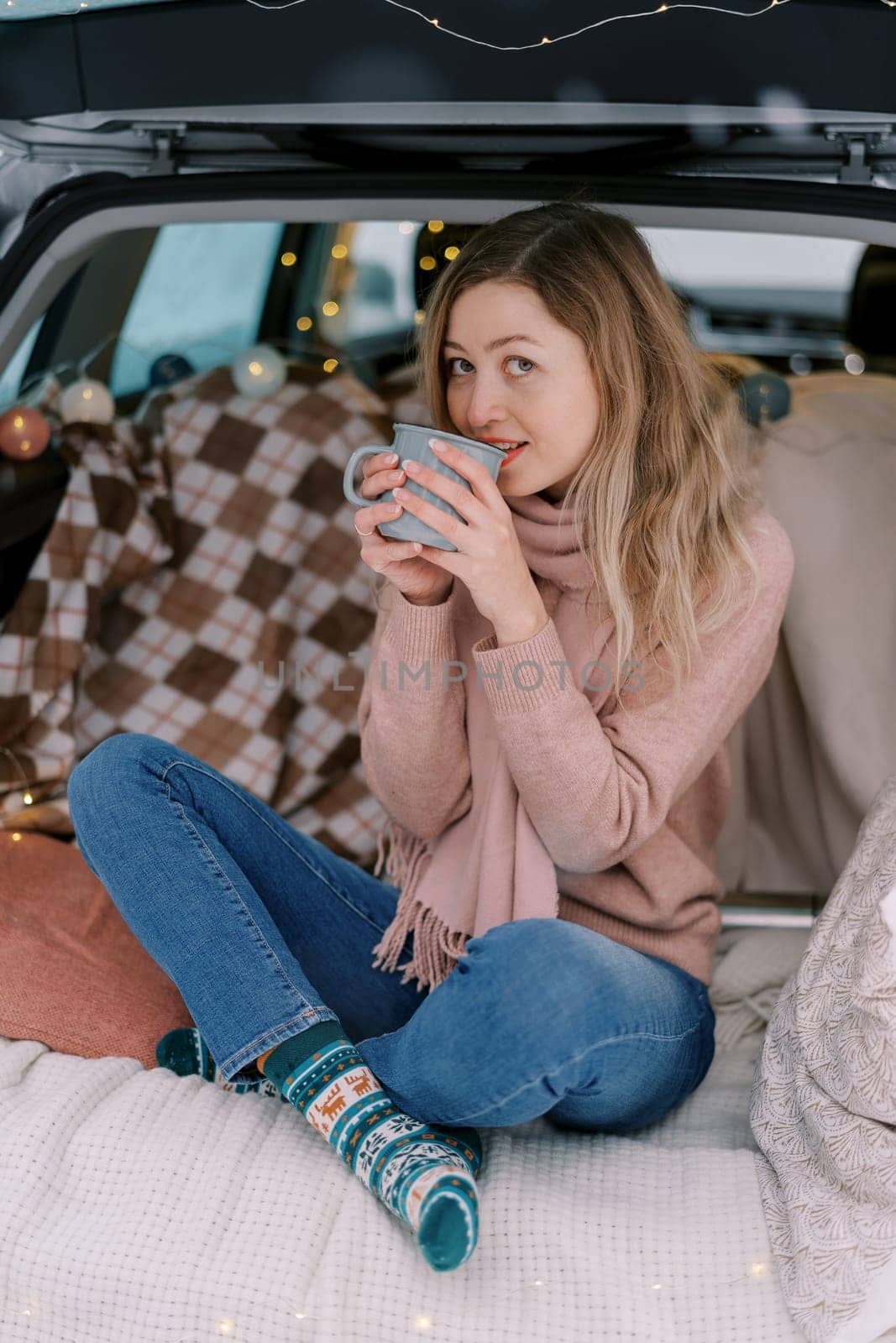 Young woman about to drink coffee while sitting in car trunk looking over cup. High quality photo