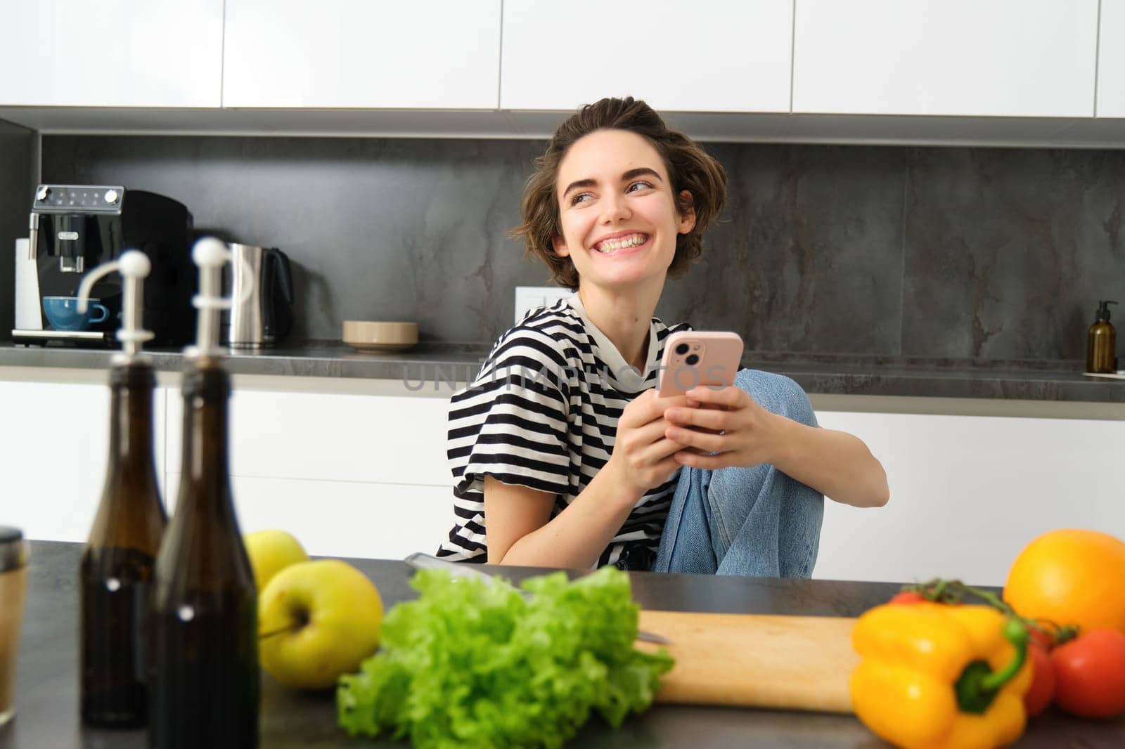 Portrait of young woman smiling, laughing and using smartphone while cooking, searching recipes on smartphone app, preparing meal from vegetables in her kitchen at home by Benzoix