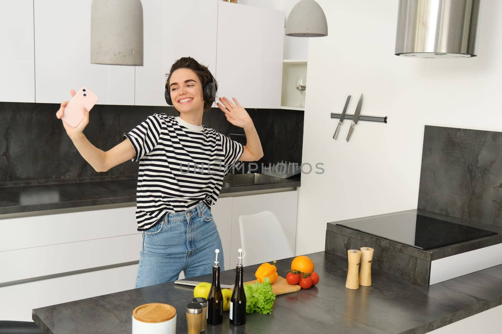 Happy woman dancing and cooking in the kitchen, listening music in earphones, making a meal, preparing salad, chopping vegetables on a counter.