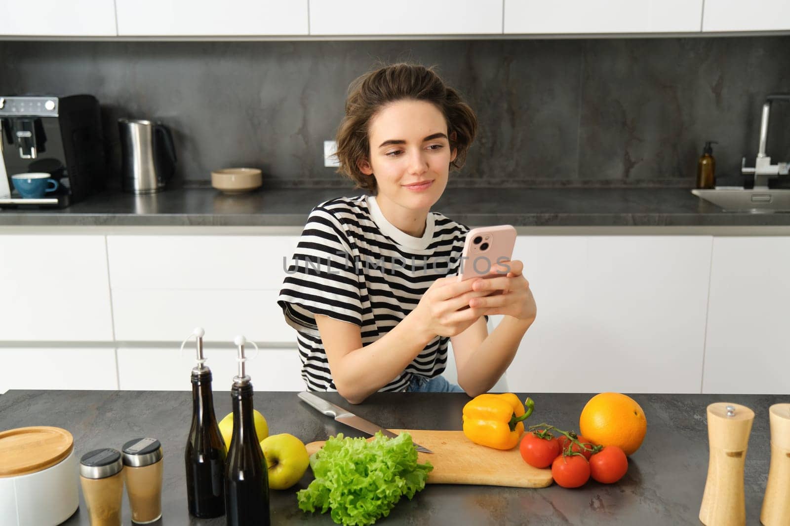 Portrait of young woman with smartphone, sitting in the kitchen and cooking salad, searching for healthy recipe online, has vegetables and chopping board on the table by Benzoix