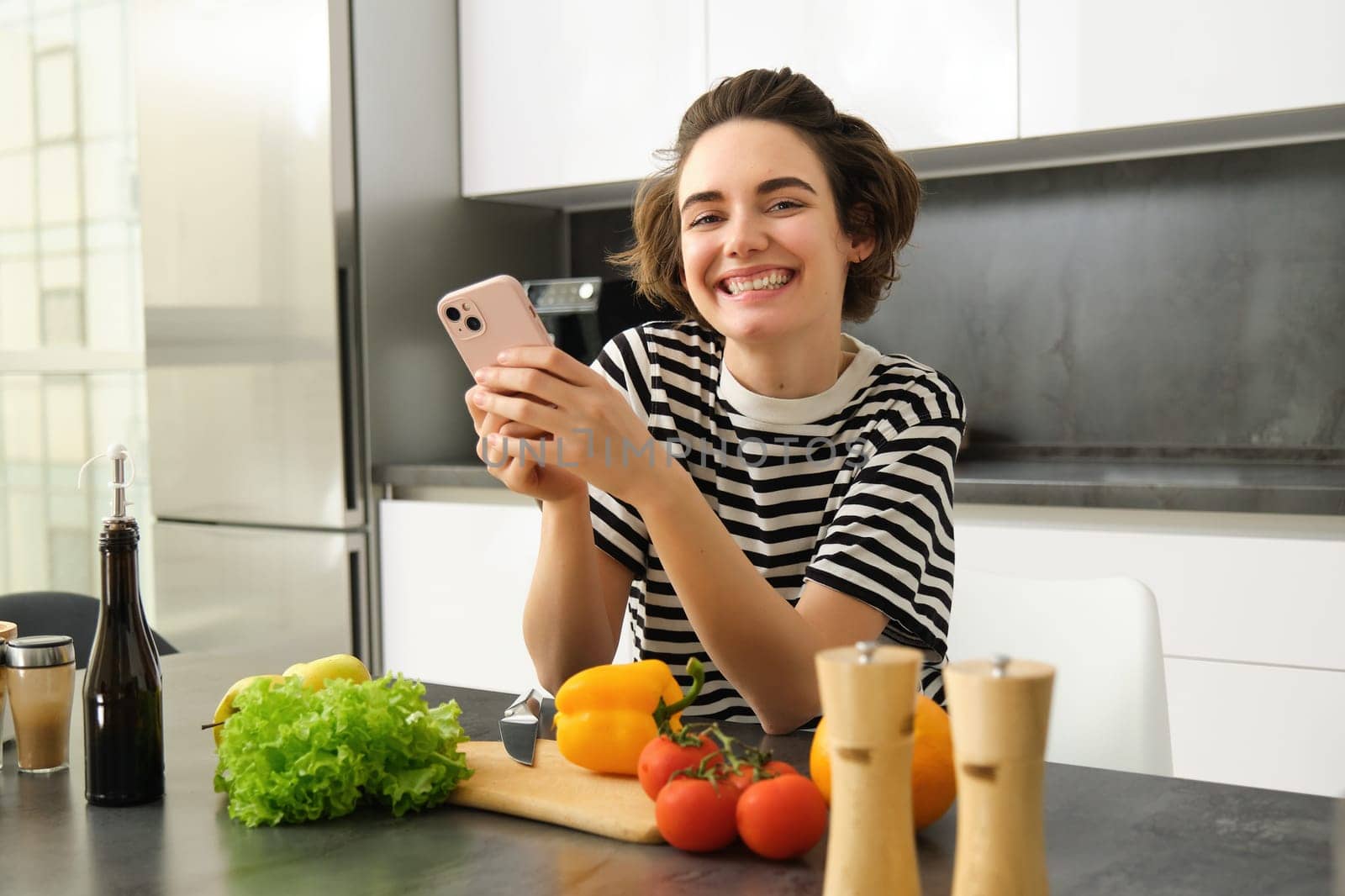 Portrait of brunette girl cooking food in the kitchen, searching recipes on social media app, holding mobile phone, standing near chopping board and vegetables, preparing healthy vegetarian meal by Benzoix