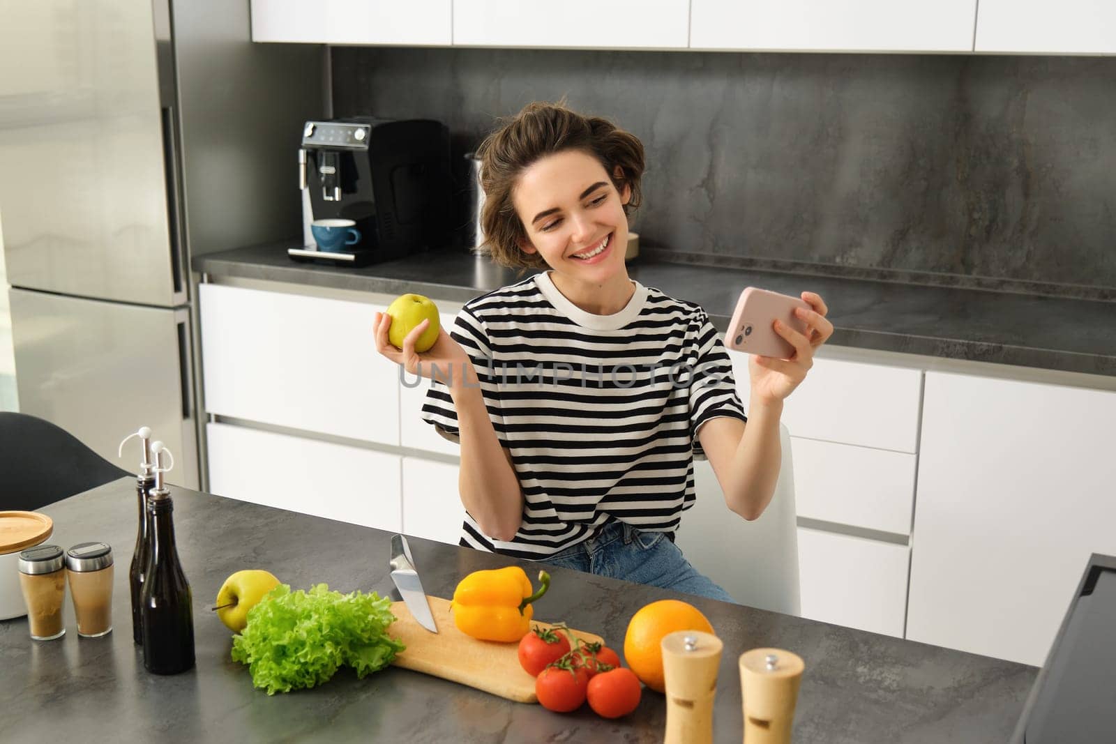 Smiling young woman, food blogger recording a video on smartphone with food, cooking and making selfie, taking pictures during meal preparation, sitting near vegetables and holding an apple by Benzoix