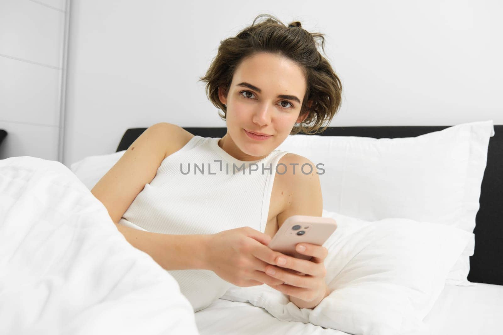 Portrait of woman lying in her bed, checking phone, sets up alarm clock to wake up in the morning.