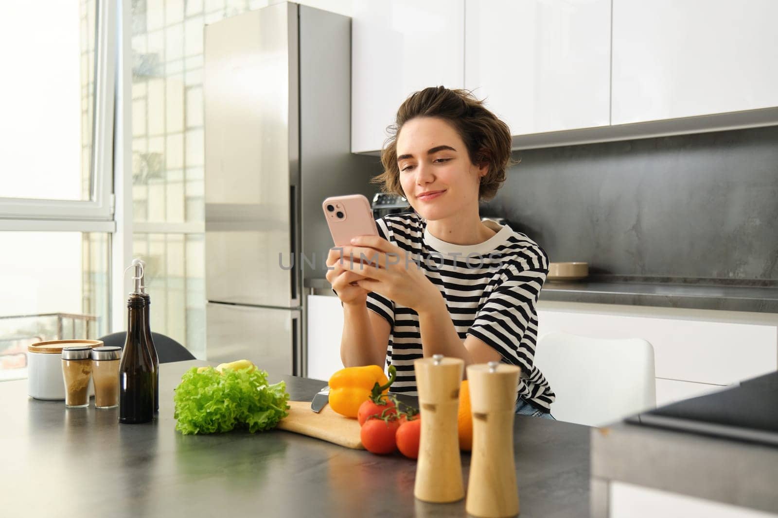 Portrait of cute modern woman cooking in the kitchen, using smartphone, reading recipe, searching meals for breakfast, leaning on counter with vegetables and chopping board.