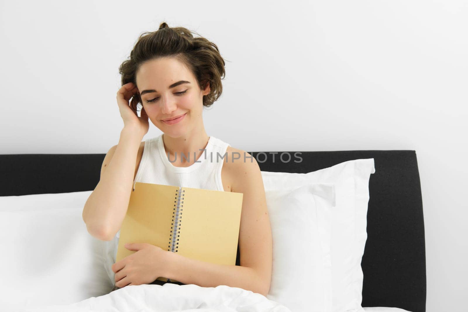 Portrait of beautiful woman in bed, sitting with her journal, writing in diary and smiling, tucking hair behind ear, reading notebook by Benzoix