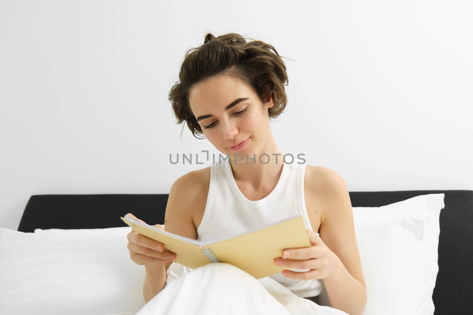 Close up portrait of young woman, reading journal, sitting in bed and studying, revising for exam before going to sleep.